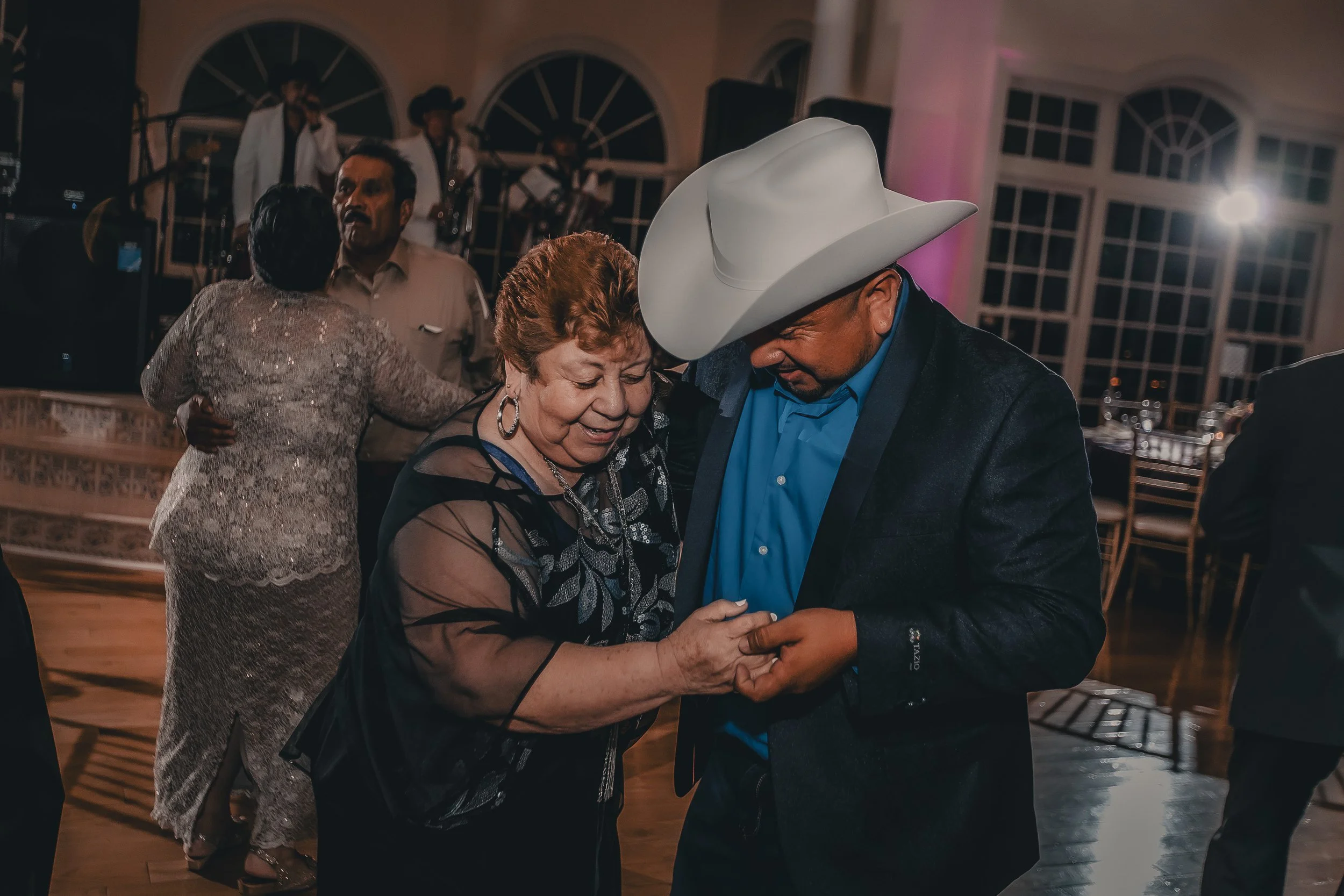 A man wearing a white cowboy hat and a blue shirt dancing with an older woman with short curly hair wearing a black top with sheer sleeves at a social event.