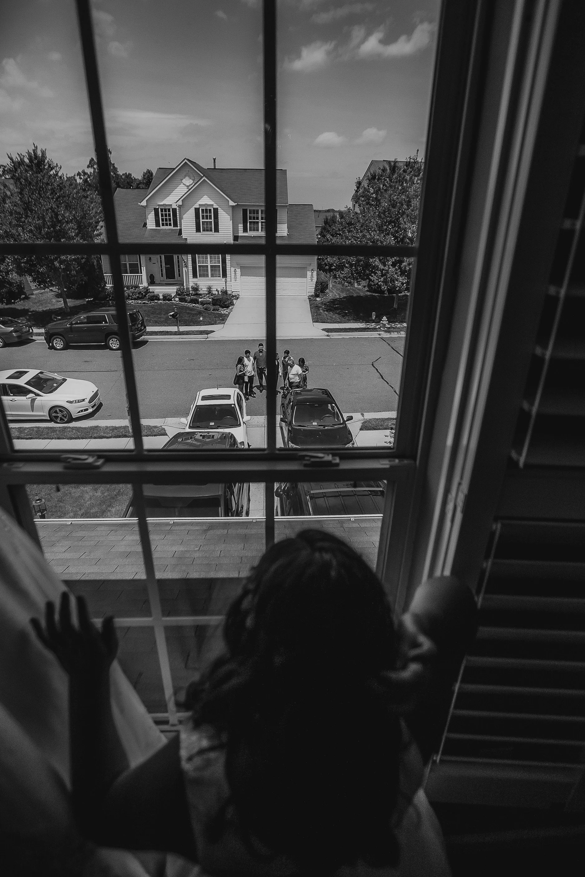 A girl looking out of a window at a group of children and adults standing on the sidewalk in front of a suburban house.