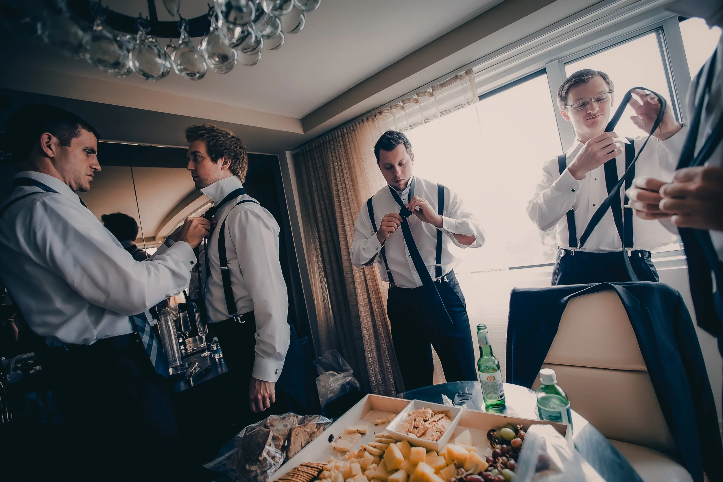 Four men in formal attire preparing for a formal event in a hotel room, with a table of snacks and drinks, including cheese, grapes, and water bottles.