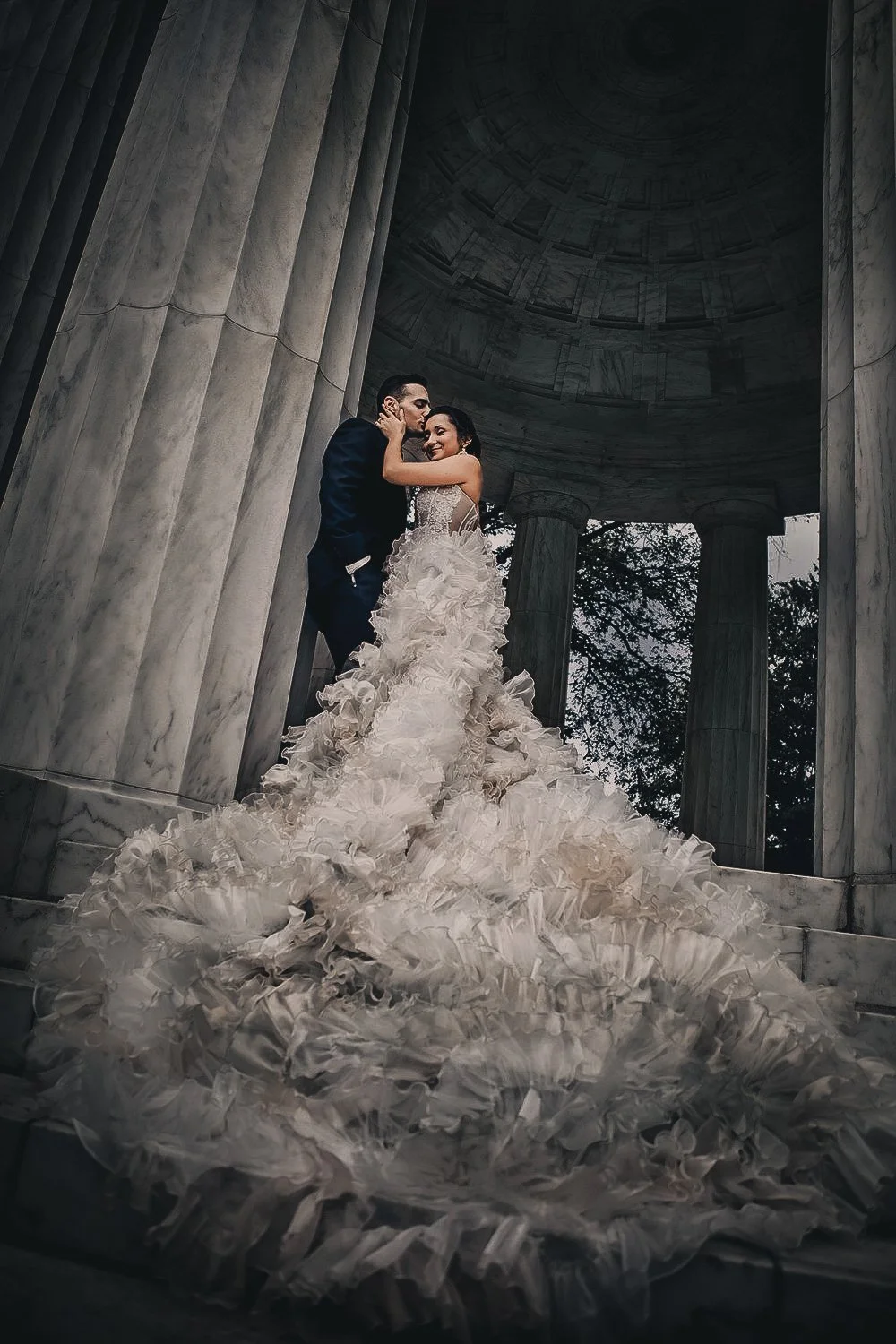 A bride and groom sharing a kiss in a marble pavilion, with the bride wearing an elaborate ruffled wedding gown and the groom in a dark suit, surrounded by classical columns and an ornate ceiling.