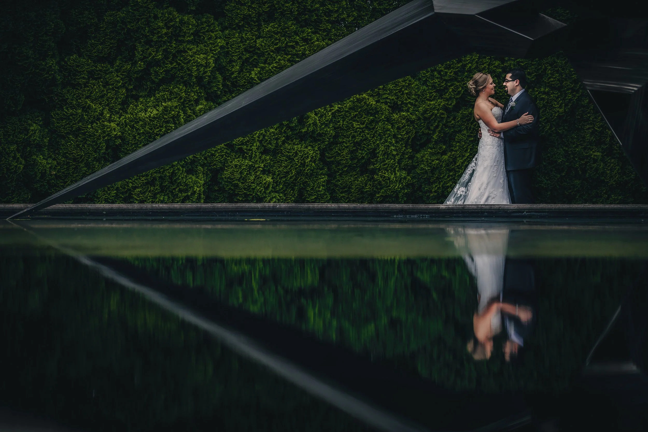 A bride and groom sharing a moment indoors, reflected in a mirror, with green foliage in the background.