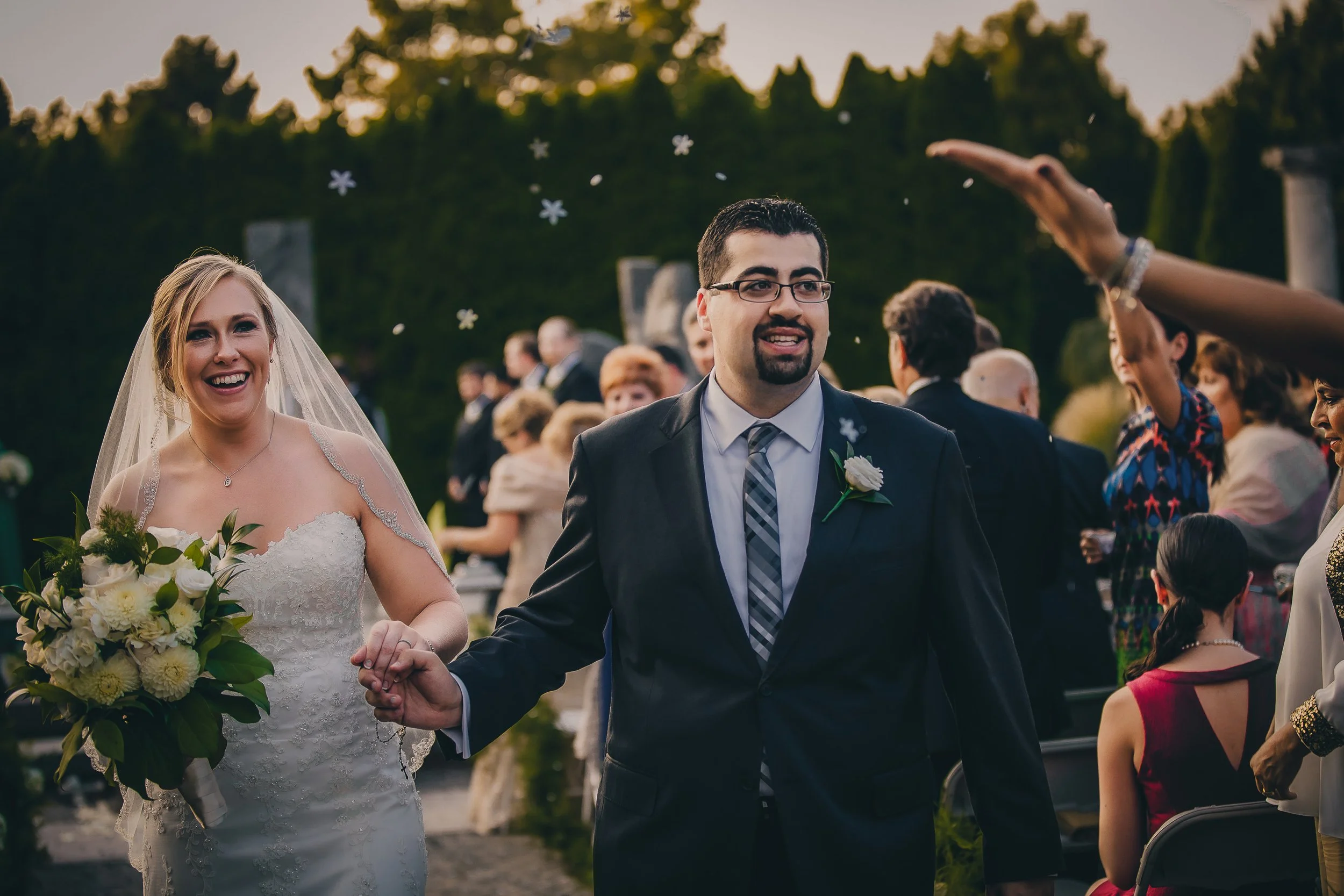 A bride and groom at their wedding ceremony outdoors, holding hands and smiling, with guests in the background celebrating.
