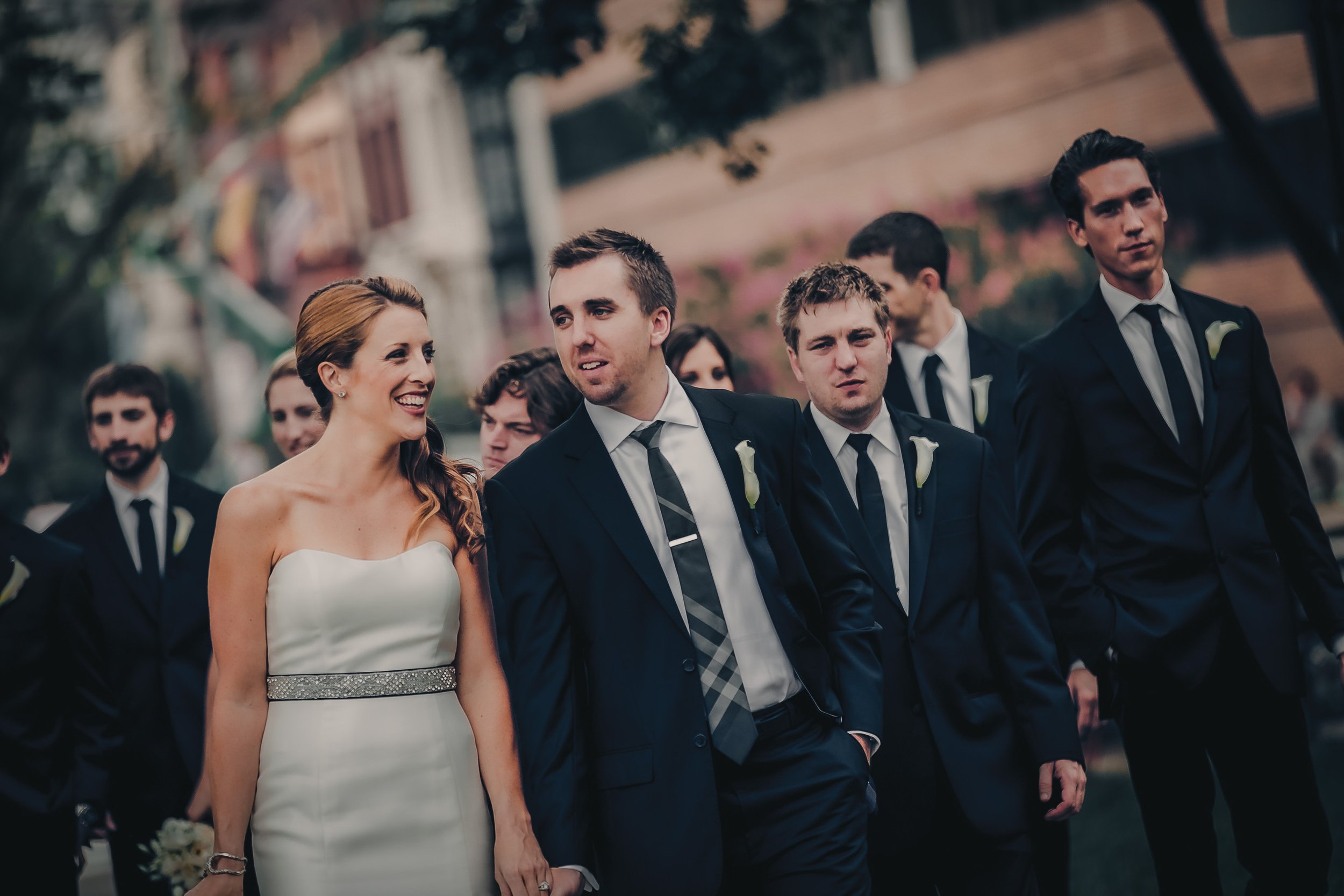 A wedding procession with a smiling bride in a white dress holding hands with a groom and walking with groomsmen dressed in black suits and white shirts outdoors.