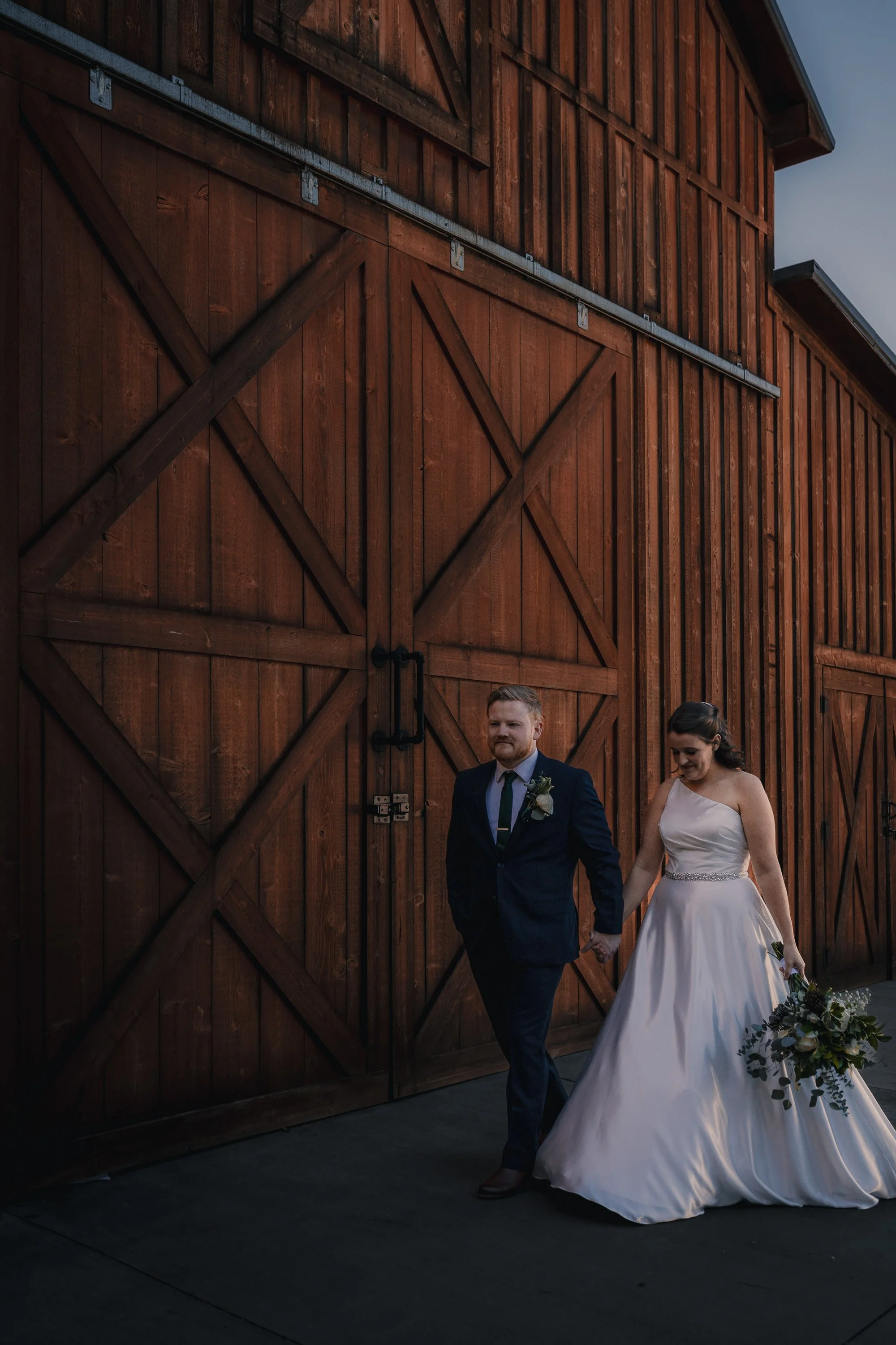 A bride and groom walking hand-in-hand outside near a large wooden barn door. The bride wears a white satin wedding dress and holds a bouquet, while the groom wears a dark suit with a boutonniere.
