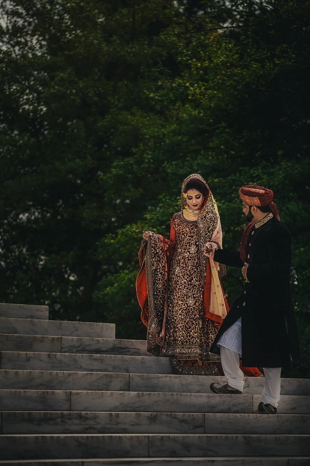 Indian bride in traditional wedding attire descending marble steps with groom in sherwani and turban at outdoor wedding.