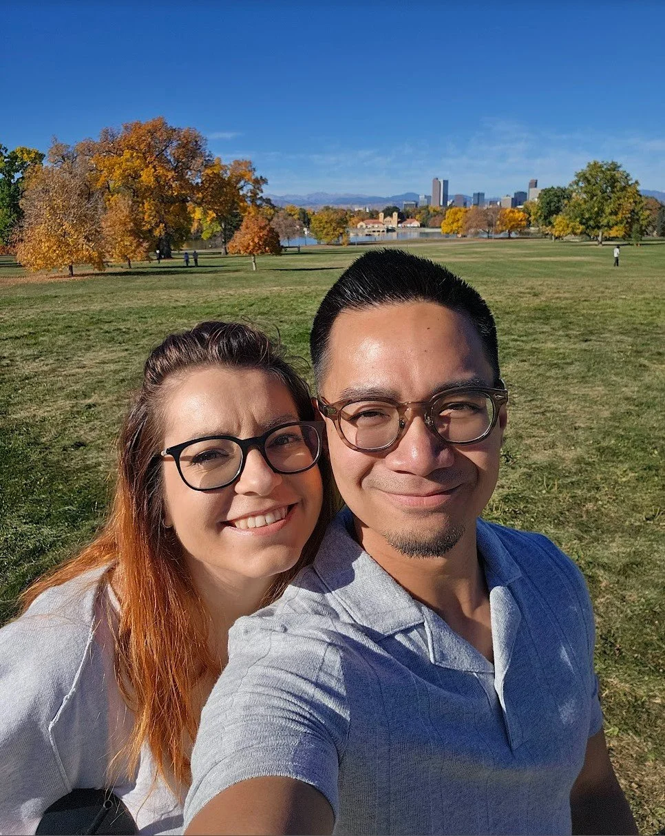 A smiling couple taking a selfie in a park with green grass, trees with autumn-colored leaves, a lake, and a city skyline in the background during daytime.