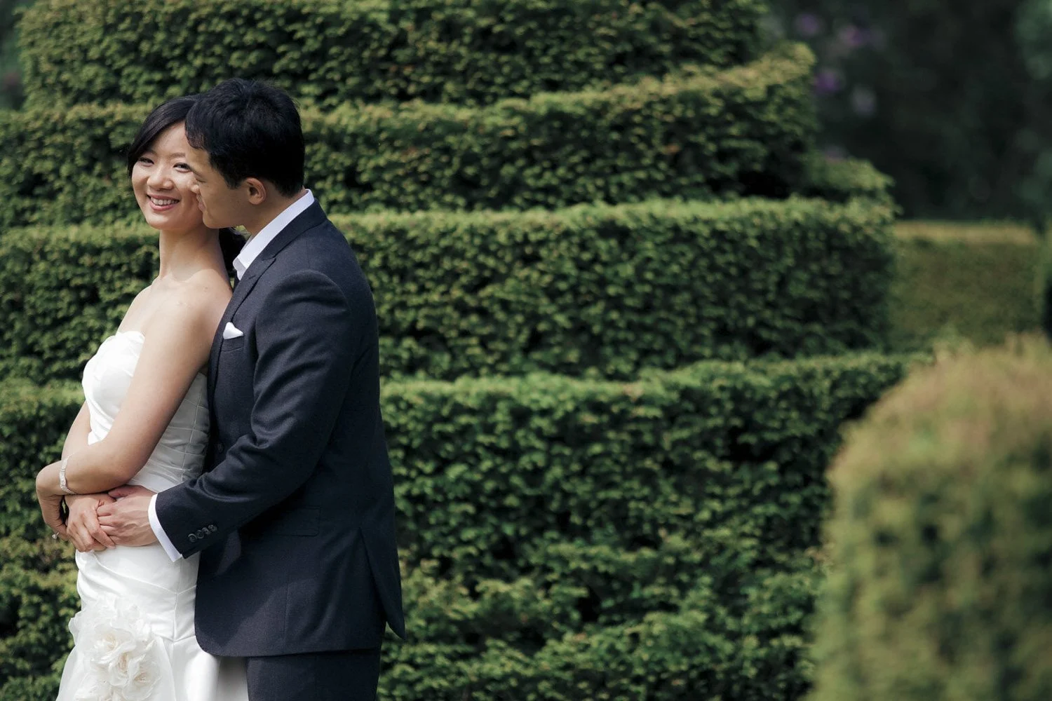 A bride and groom embracing against the backdrop of a large trimmed bush.