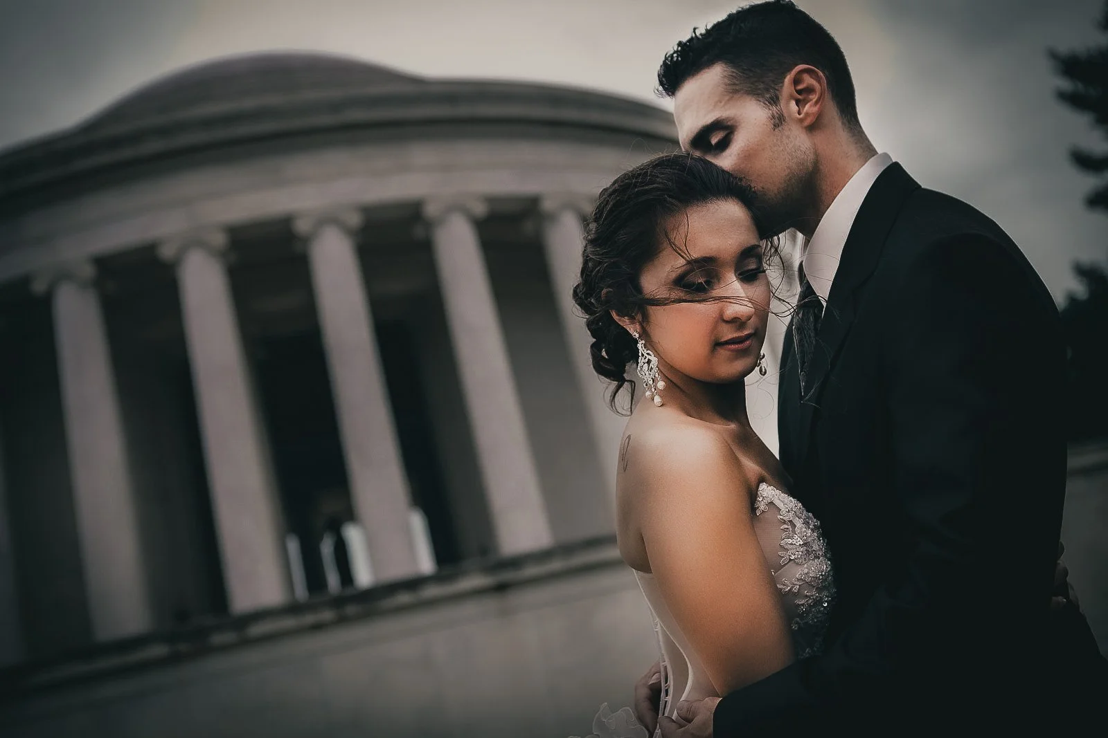 A couple dressed in wedding attire, with the bride in a lace wedding gown and the groom in a black tuxedo, embracing in front of a building with tall columns on a cloudy day.