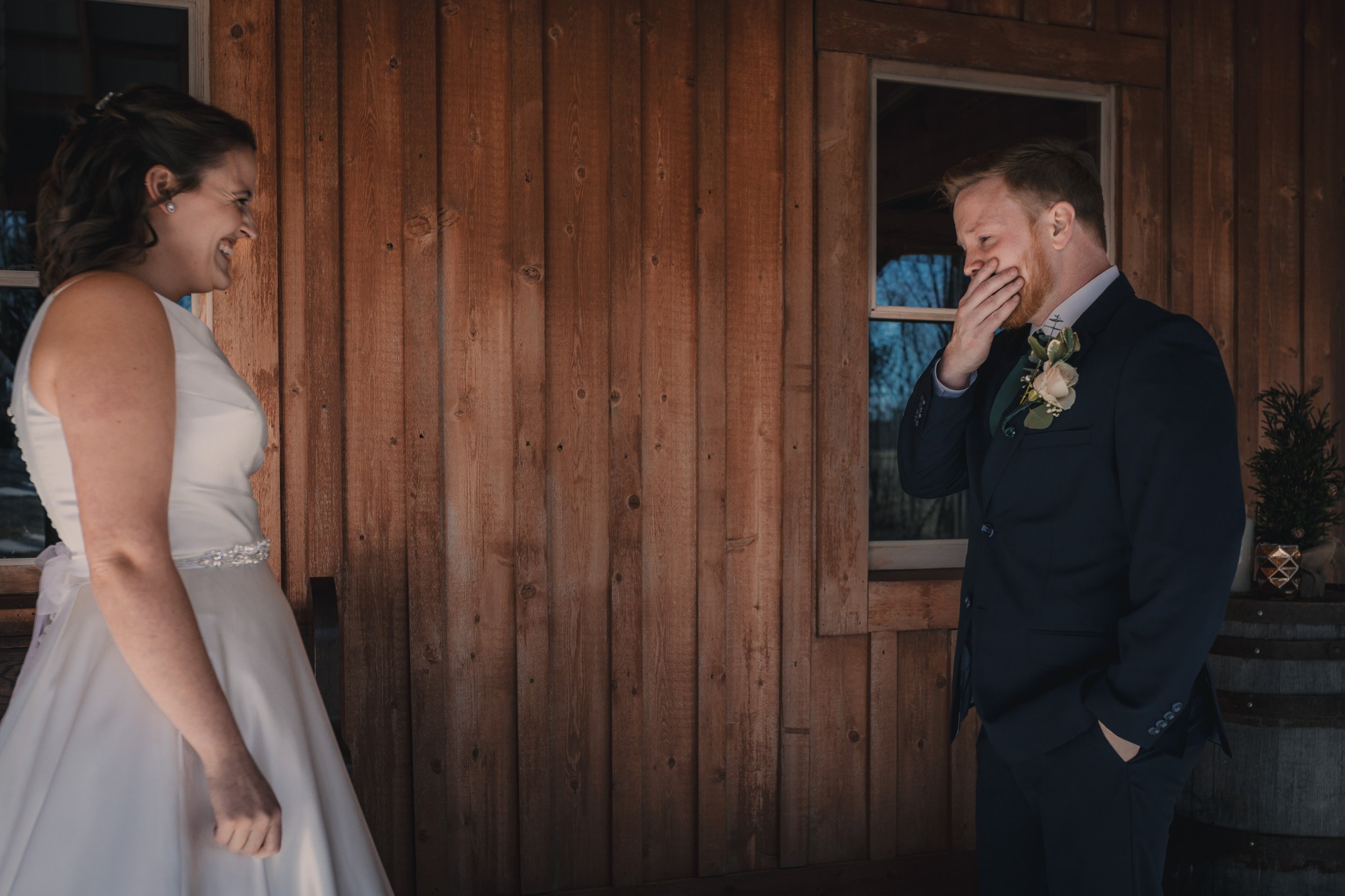 A bride and groom are smiling and laughing during their wedding in front of a wooden wall, with the bride on the left wearing a white dress and the groom on the right in a black suit with a boutonniere.