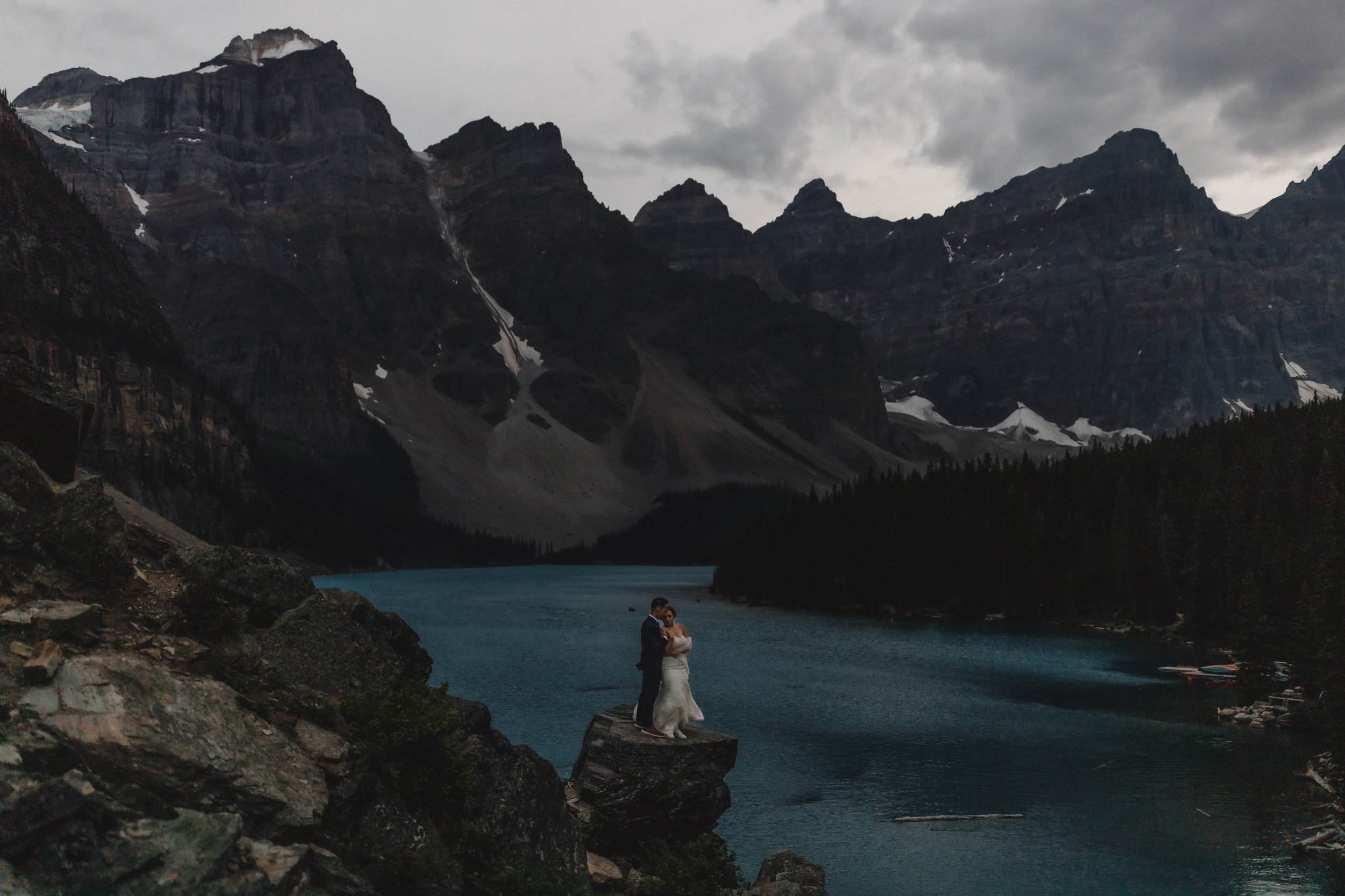 A couple standing on a rock by a lake, surrounded by mountains and trees, under a cloudy sky.