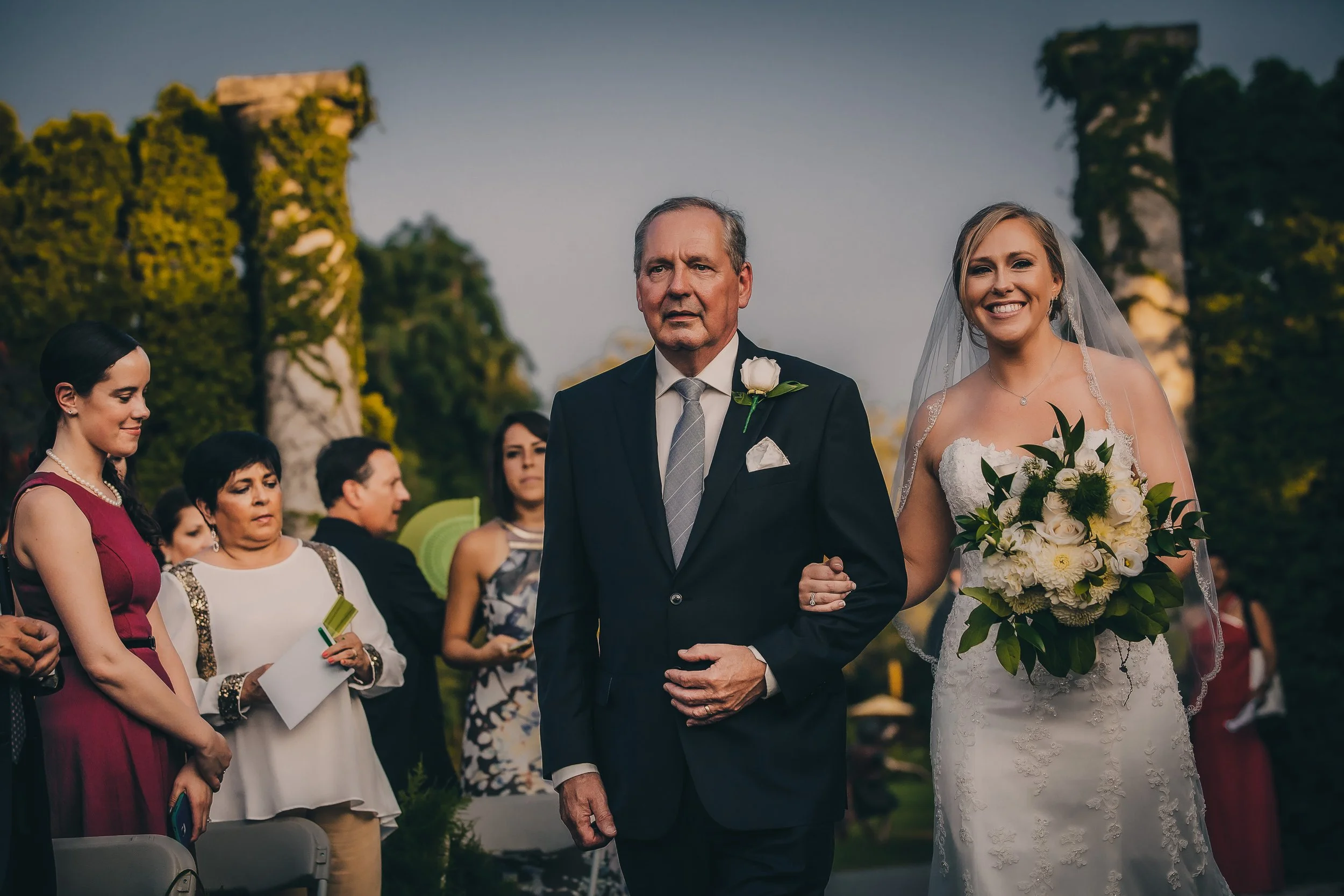 A bride in a white wedding dress holding a bouquet and walking down the aisle, accompanied by an older man in a dark suit, at an outdoor wedding ceremony with guests in the background.