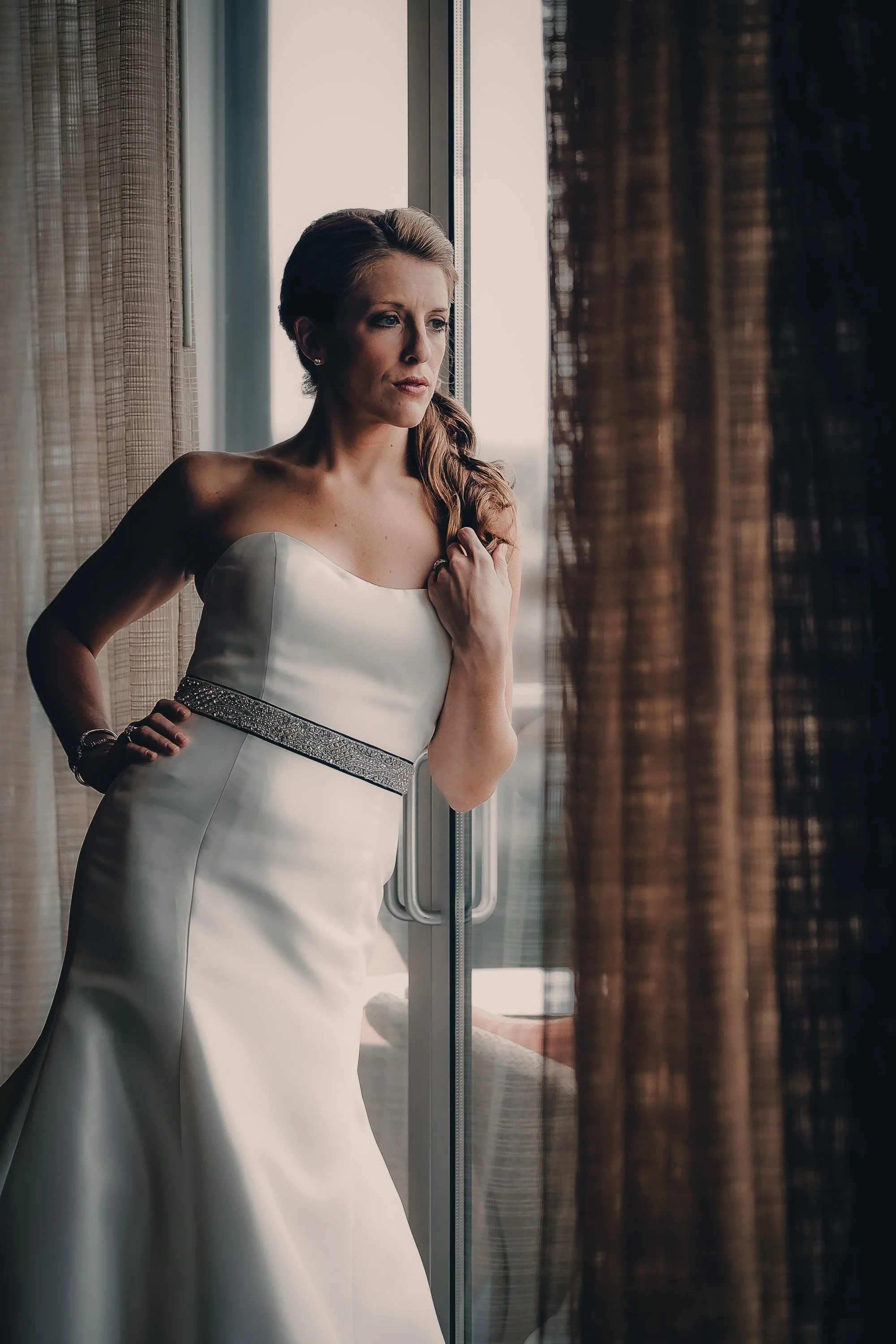 A woman in a white wedding dress, standing near a window, gazing outside thoughtfully.
