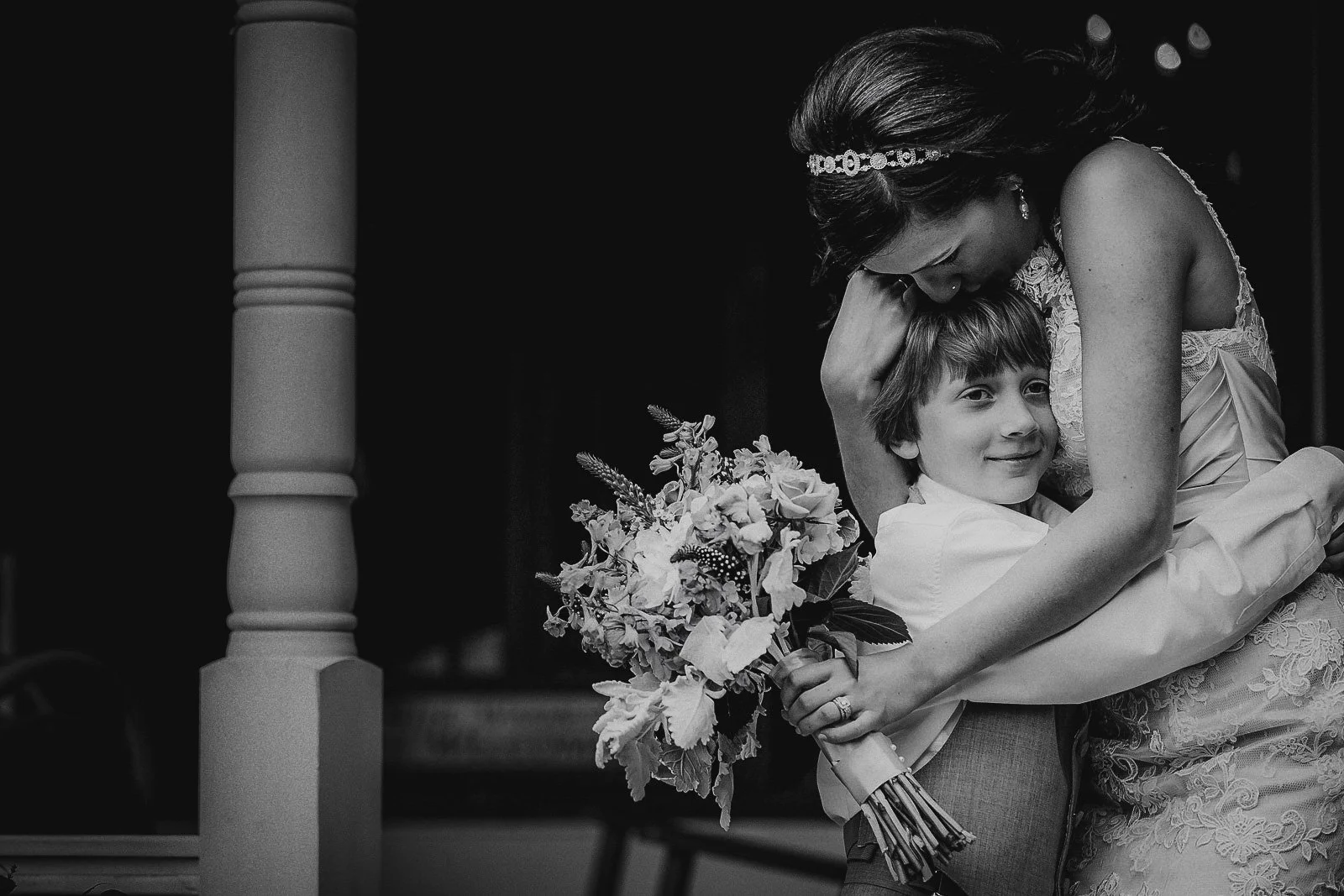 A woman in a wedding dress hugging a young boy holding a bouquet of flowers, smiling and leaning into her affection, black and white photo.