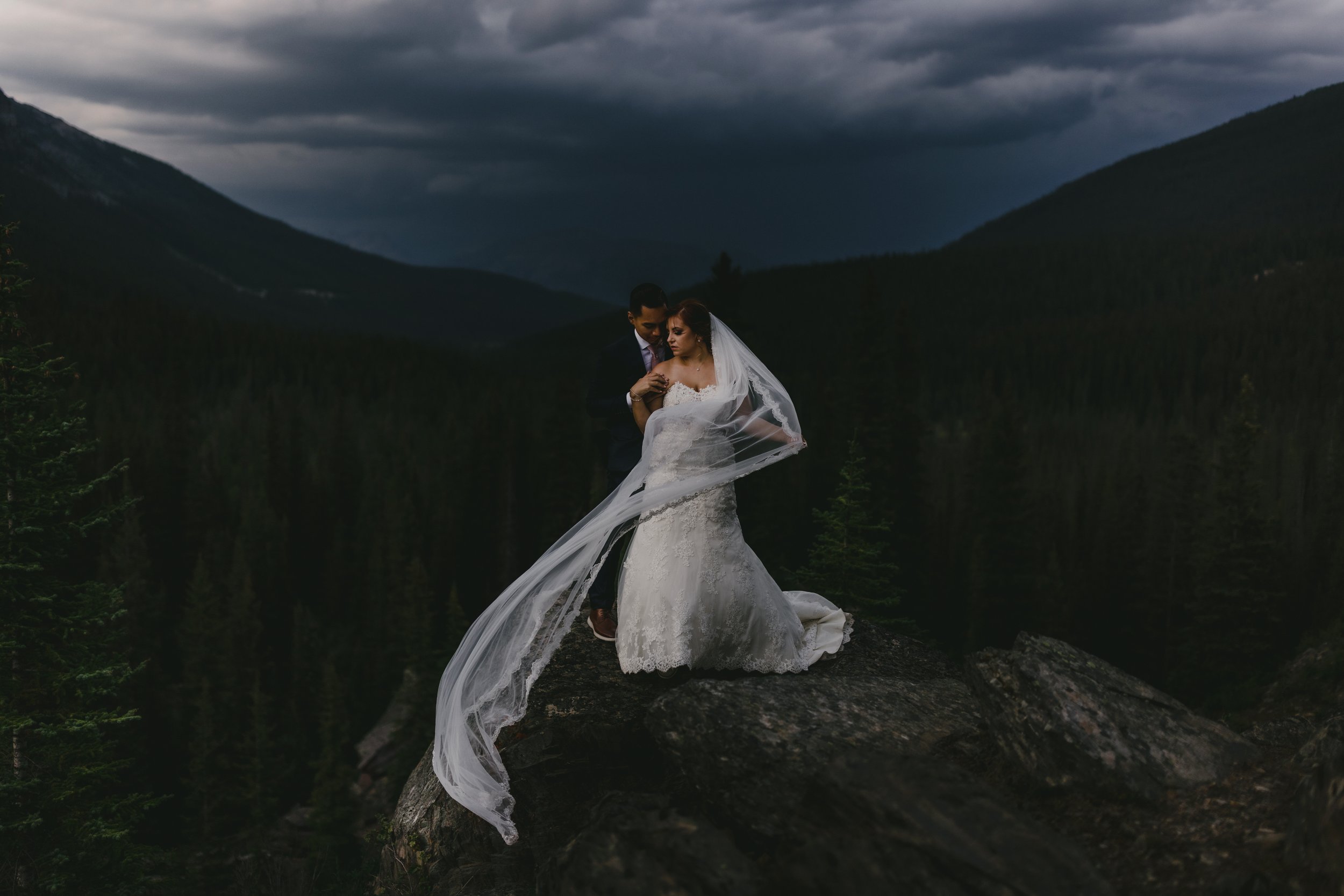 A bride and groom standing on a rock during a stormy evening with dark clouds and mountains in the background.