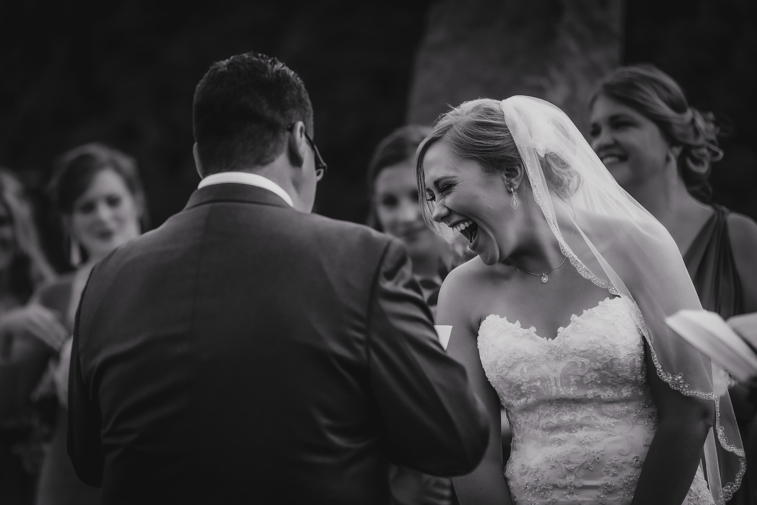 A black and white photo of a wedding ceremony showing a bride laughing with her eyes closed and a groom with his back to the camera, surrounded by bridesmaids.