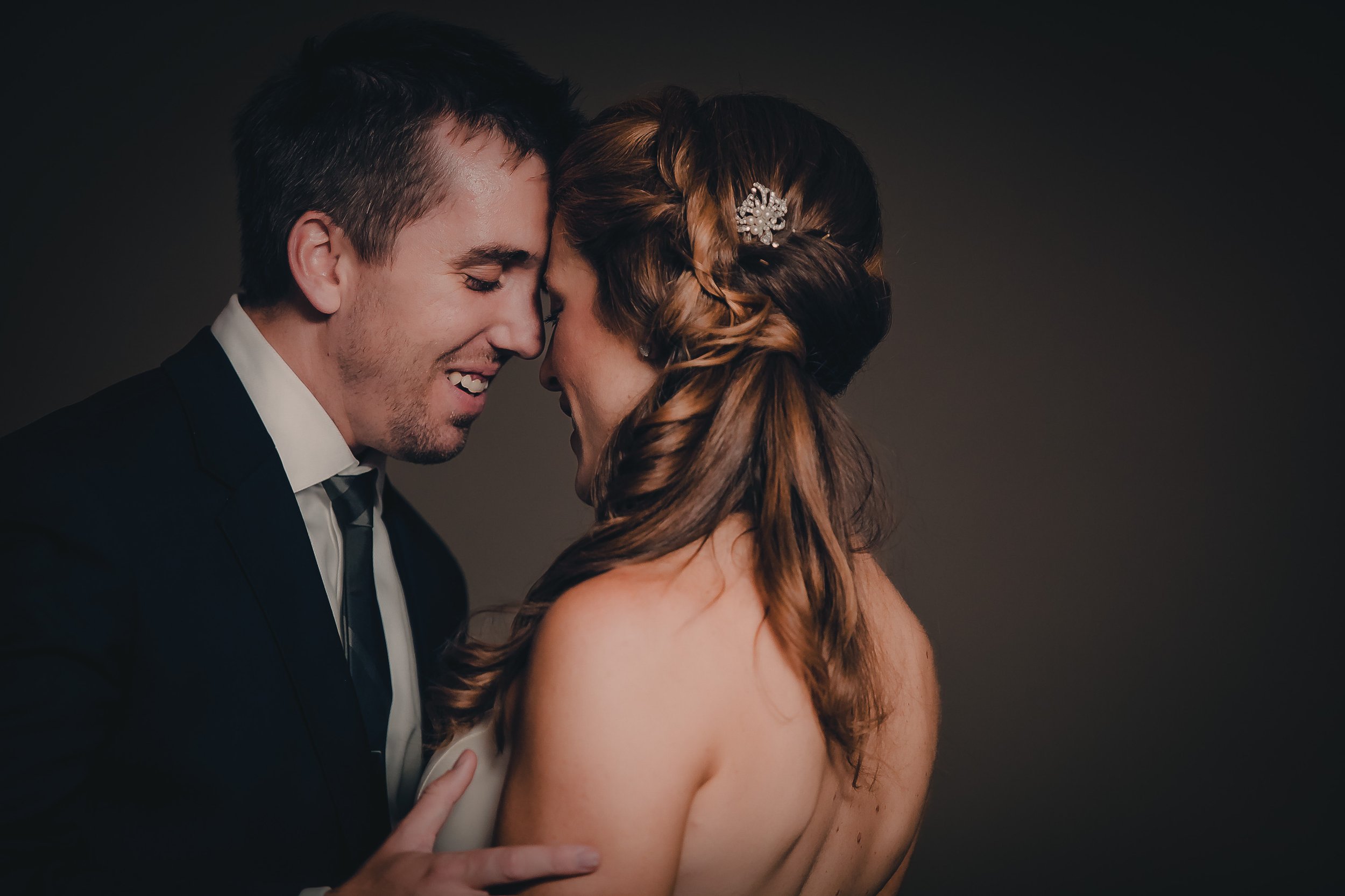 A couple in wedding attire smiling and leaning their foreheads together against a dark background.