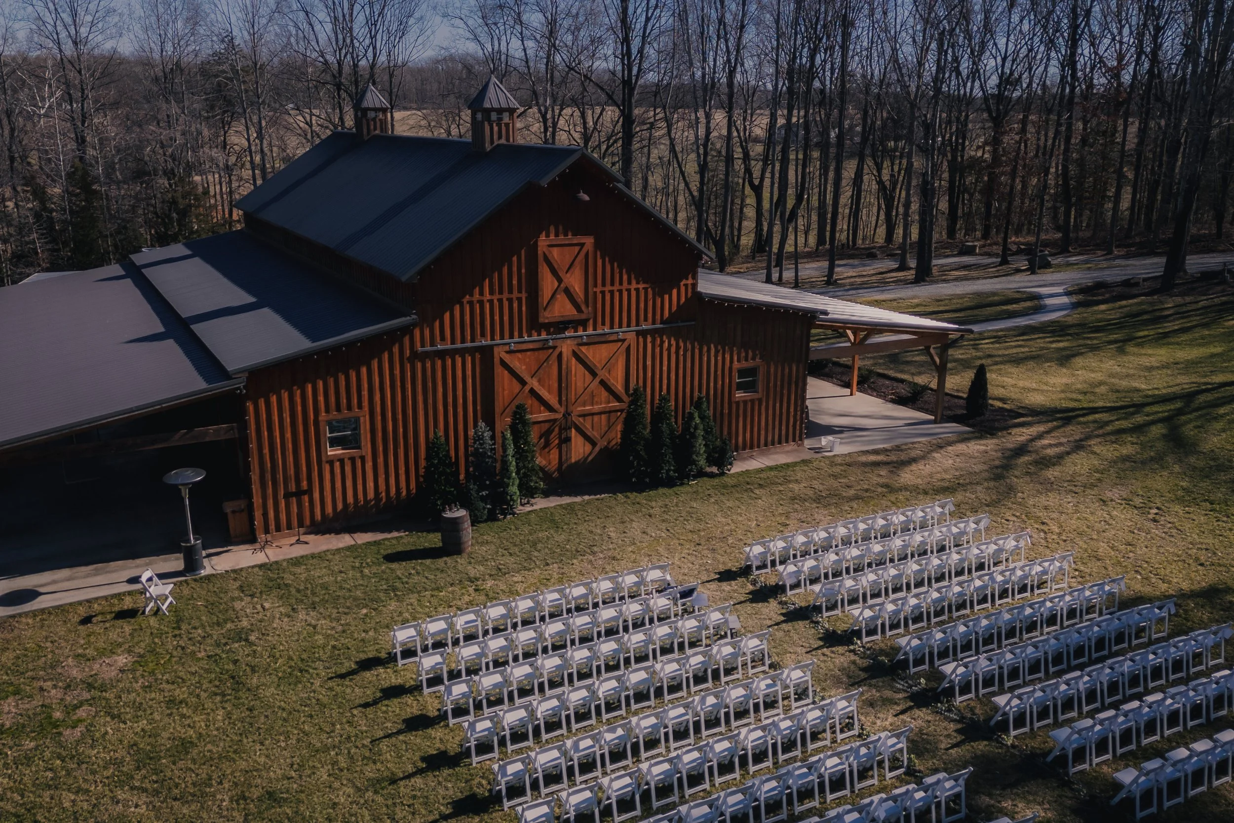 An outdoor wedding setup with rows of white chairs facing a rustic wooden barn, with greenery and leafless trees in the background.
