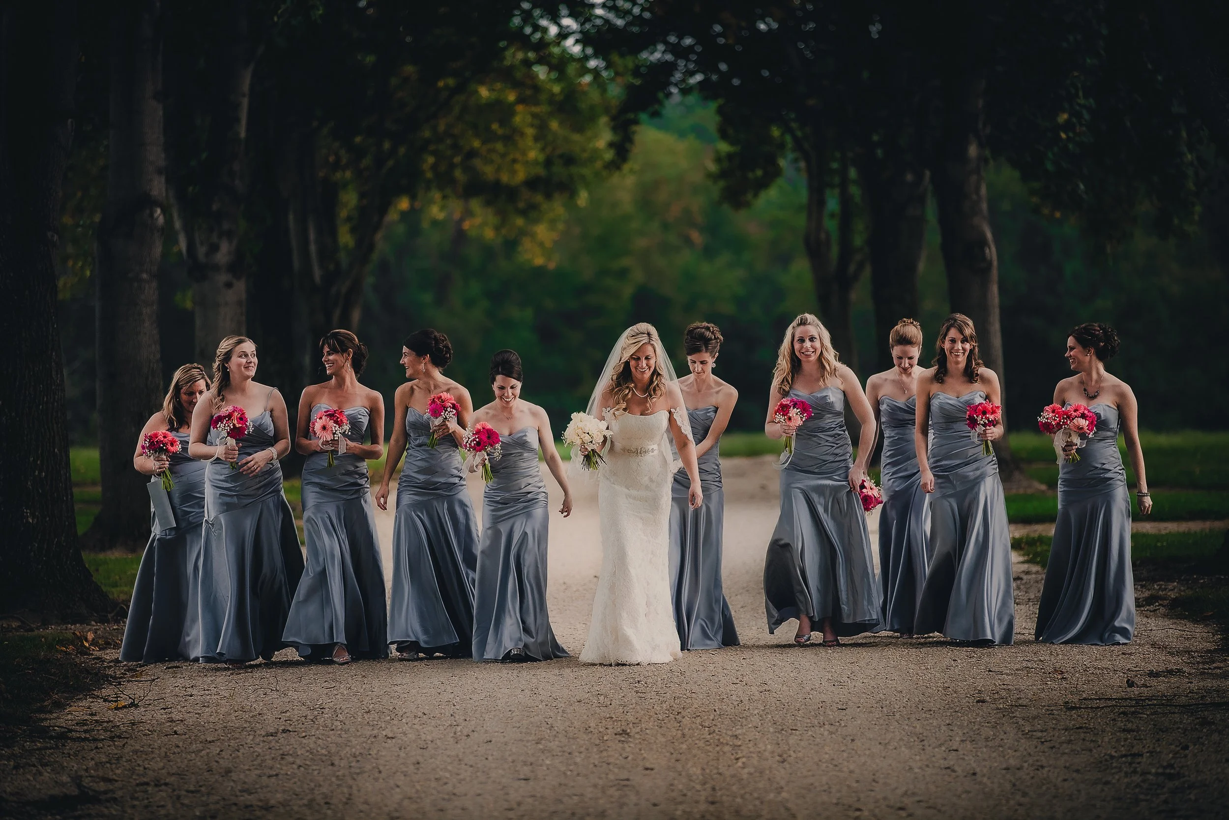 A bride in a white wedding dress walking with her bridesmaids in matching gray dresses through a wooded area, each holding a bouquet of pink and white flowers.