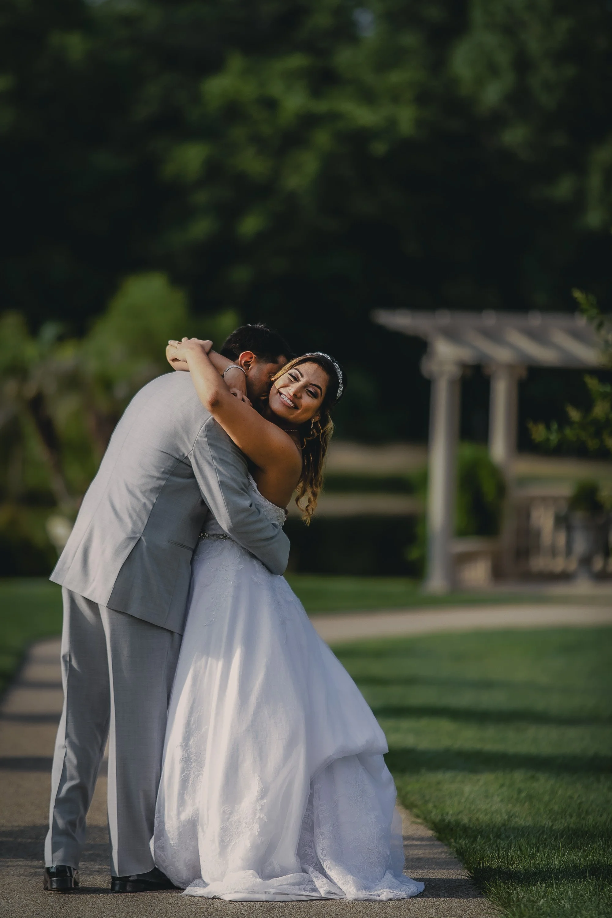 A newlywed couple hugging outdoors on a sunny day, with the bride in a white wedding dress and the groom in a gray suit, standing on a pathway with green trees and a pavilion in the background.