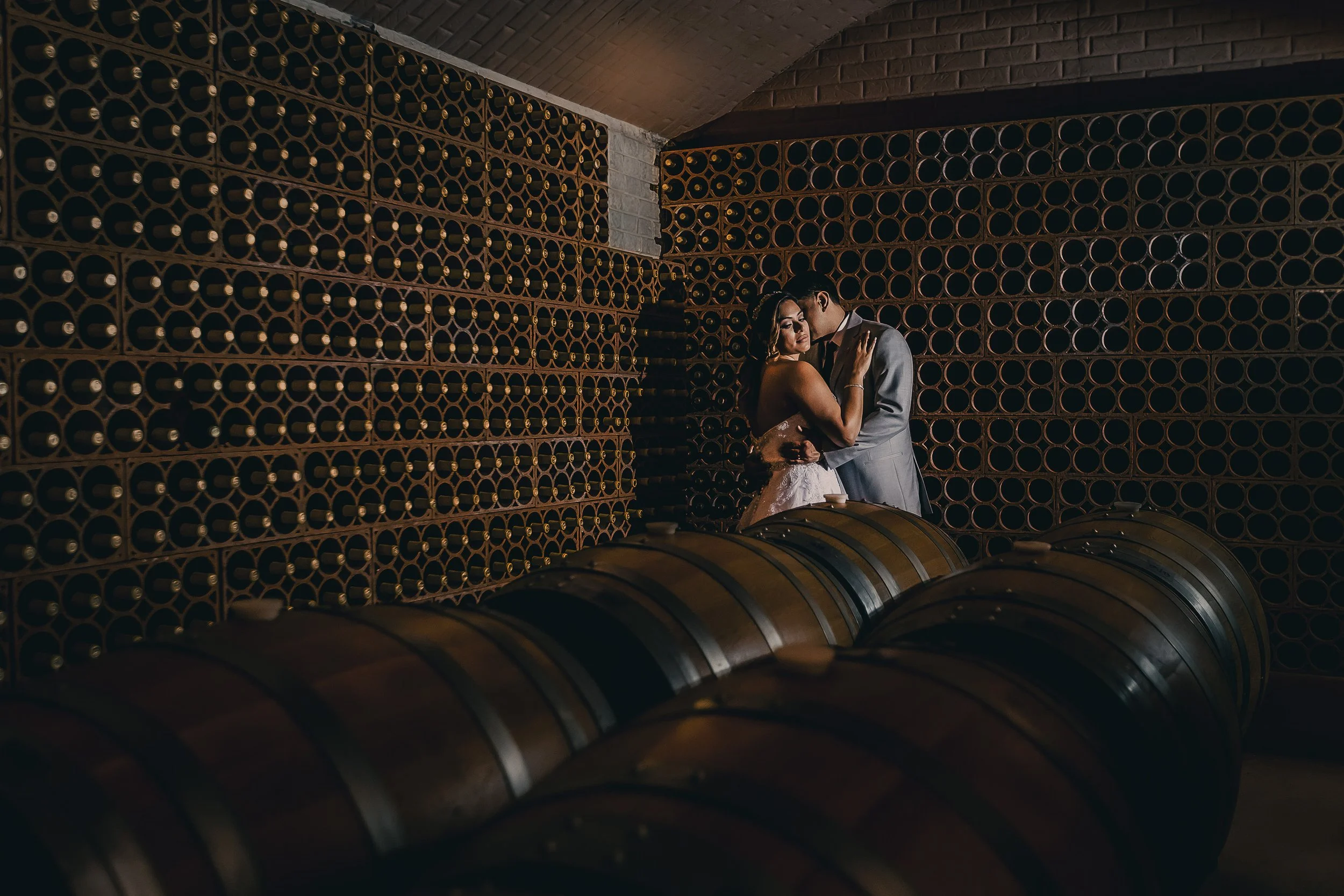 A bride and groom embrace in a cellar filled with wine barrels and wine racks.