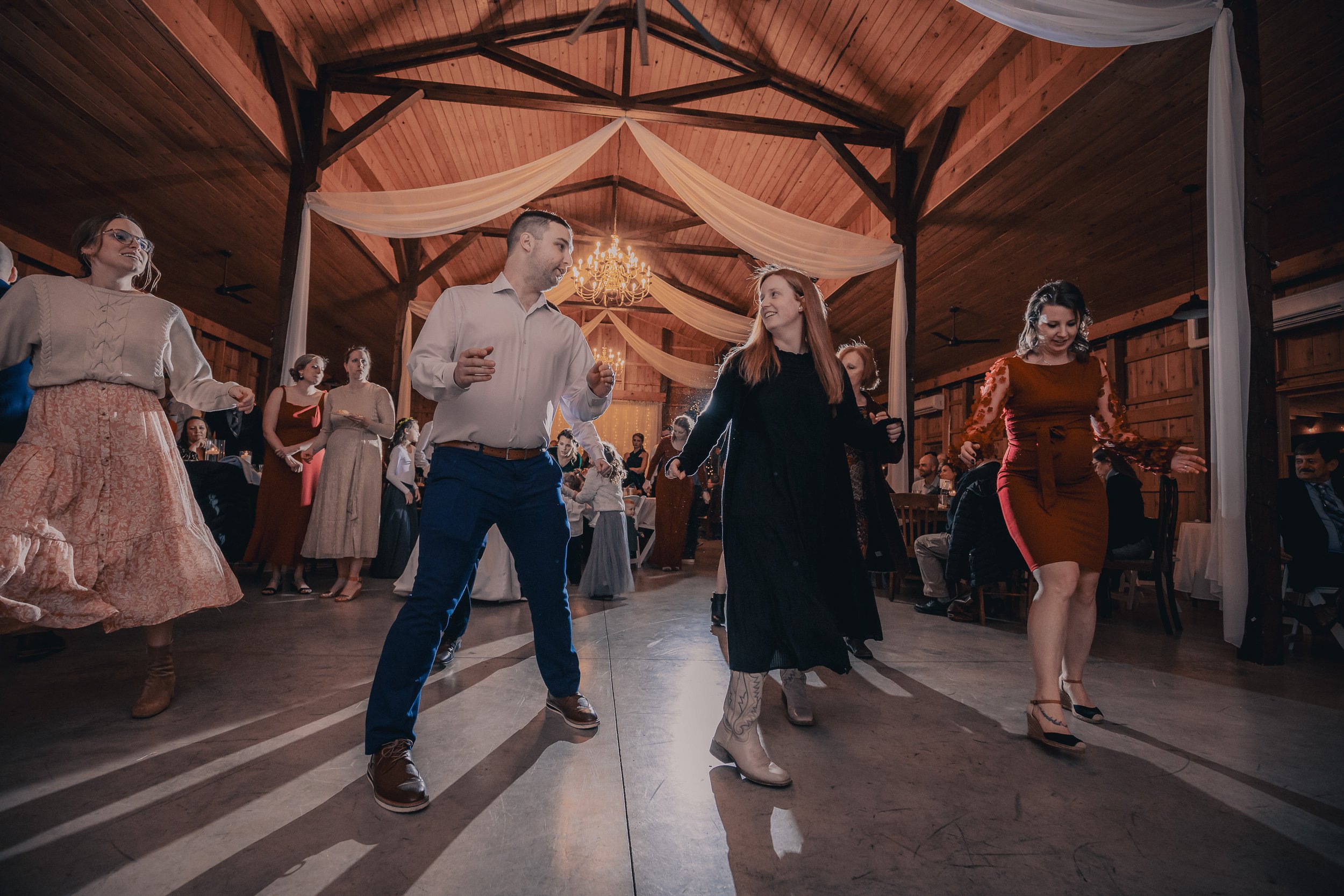 People dancing at a wedding reception in a rustic barn, with a wooden ceiling, chandeliers, and white drapes.
