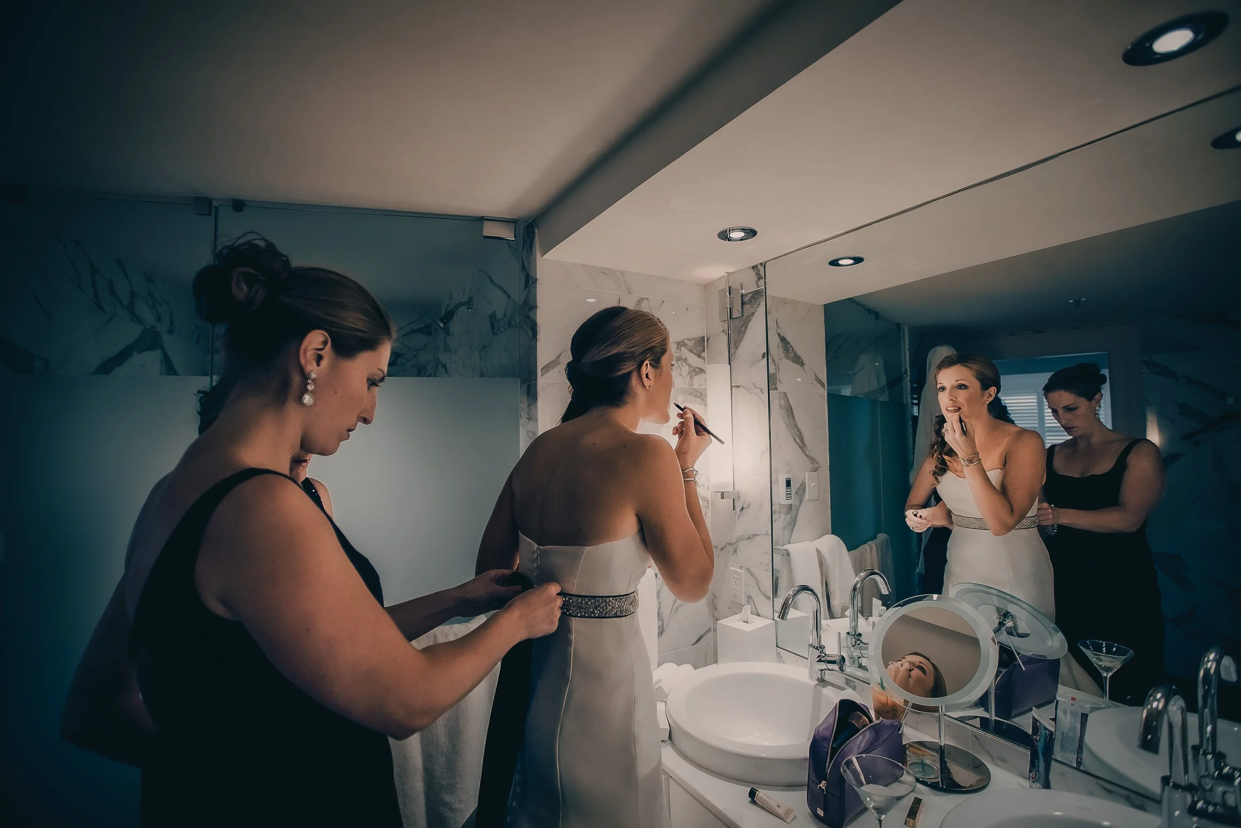 Three women preparing for a wedding in a bathroom, two women are helping the bride, who is wearing a white dress with a jeweled belt, while the third woman adjusts something on her dress. The bride is looking into the mirror, and the bathroom has mar