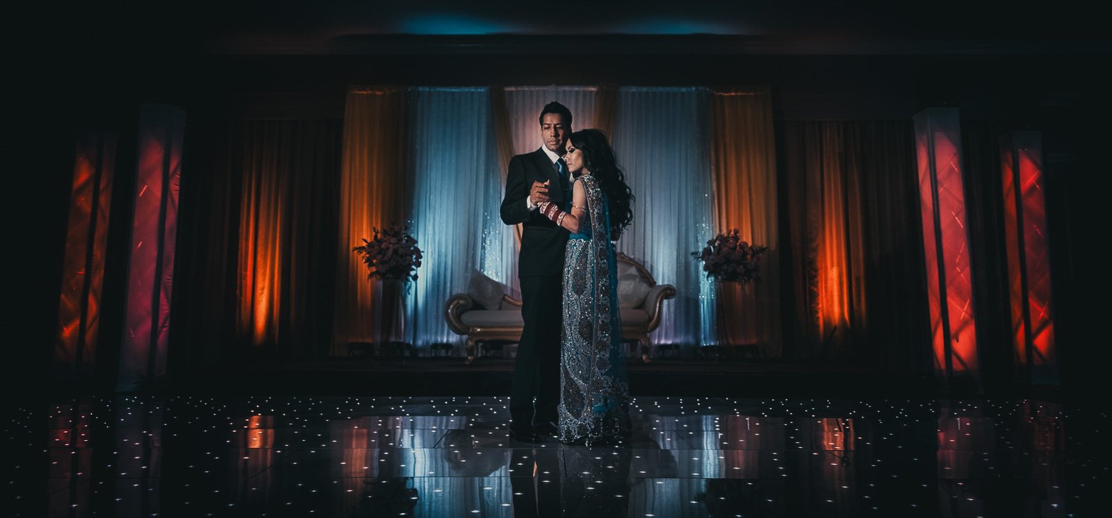 A man and woman dancing closely on a stage with colorful drapes and flowers, illuminated by soft lighting.