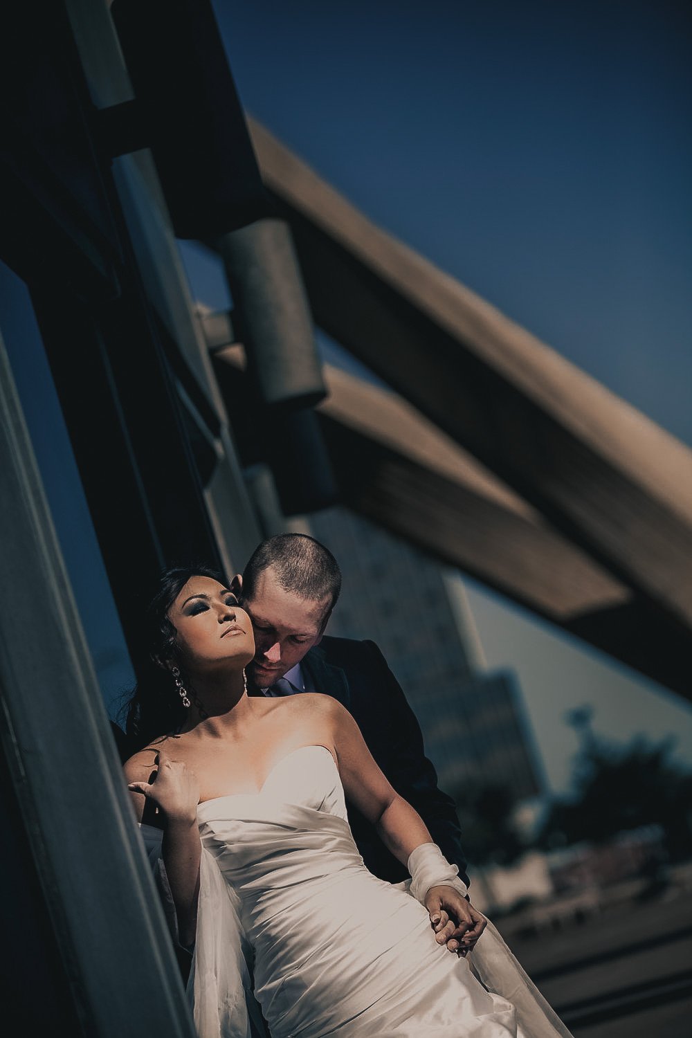 A bride in a white wedding dress with dark hair and earrings, and a groom in a dark suit, standing close together under a bridge with city buildings in the background during evening or night.