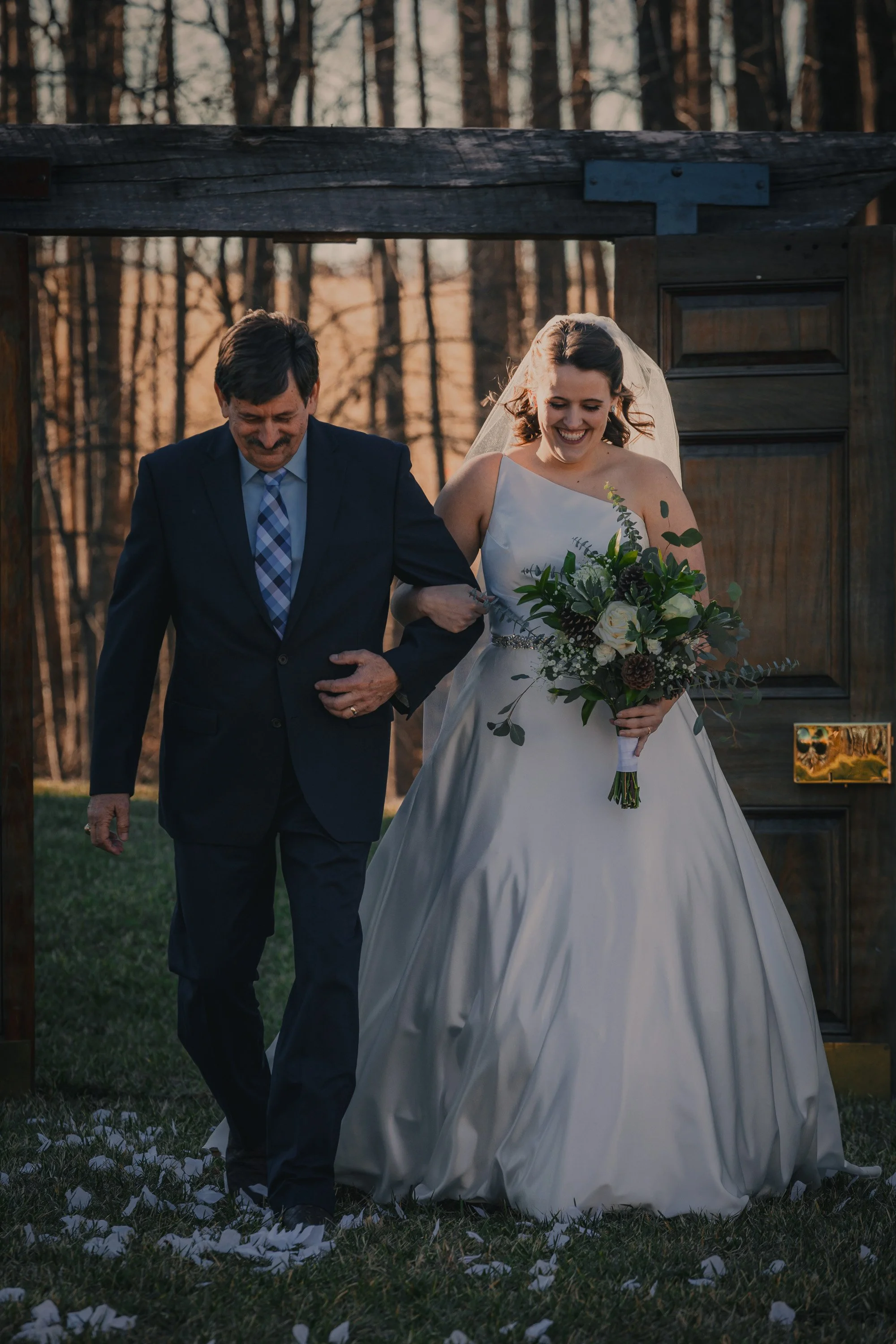 A bride in a white wedding gown holding a bouquet of flowers walking with an older man in a suit, outdoors during sunset, in front of a wooden gate.