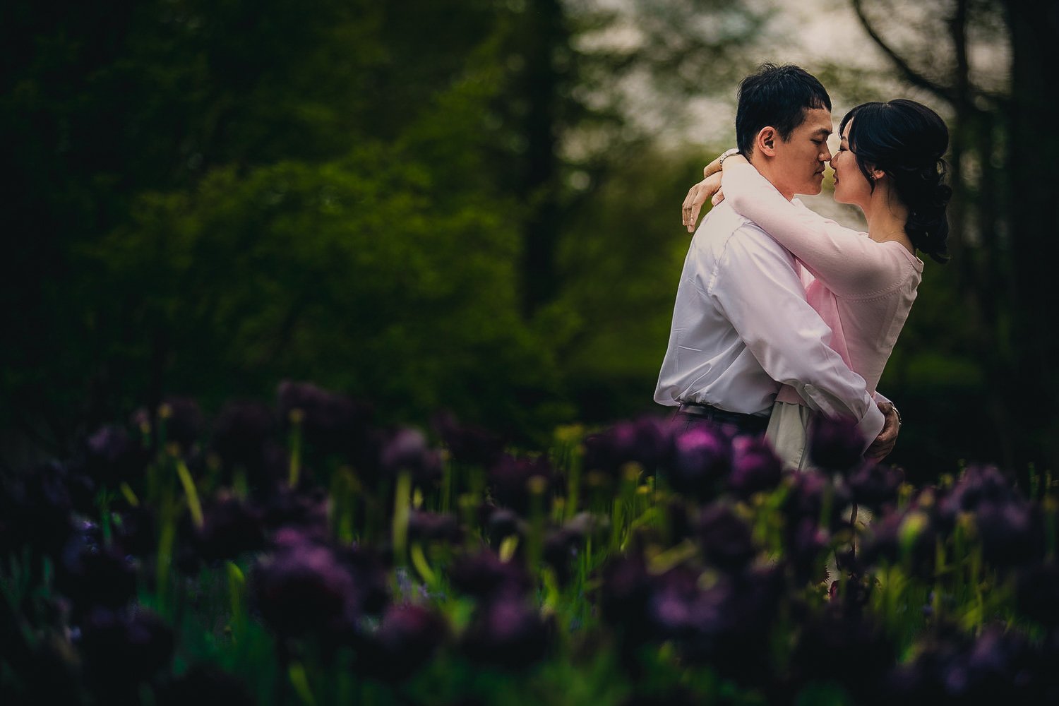A couple embracing in a garden with purple flowers in the foreground and green trees in the background.