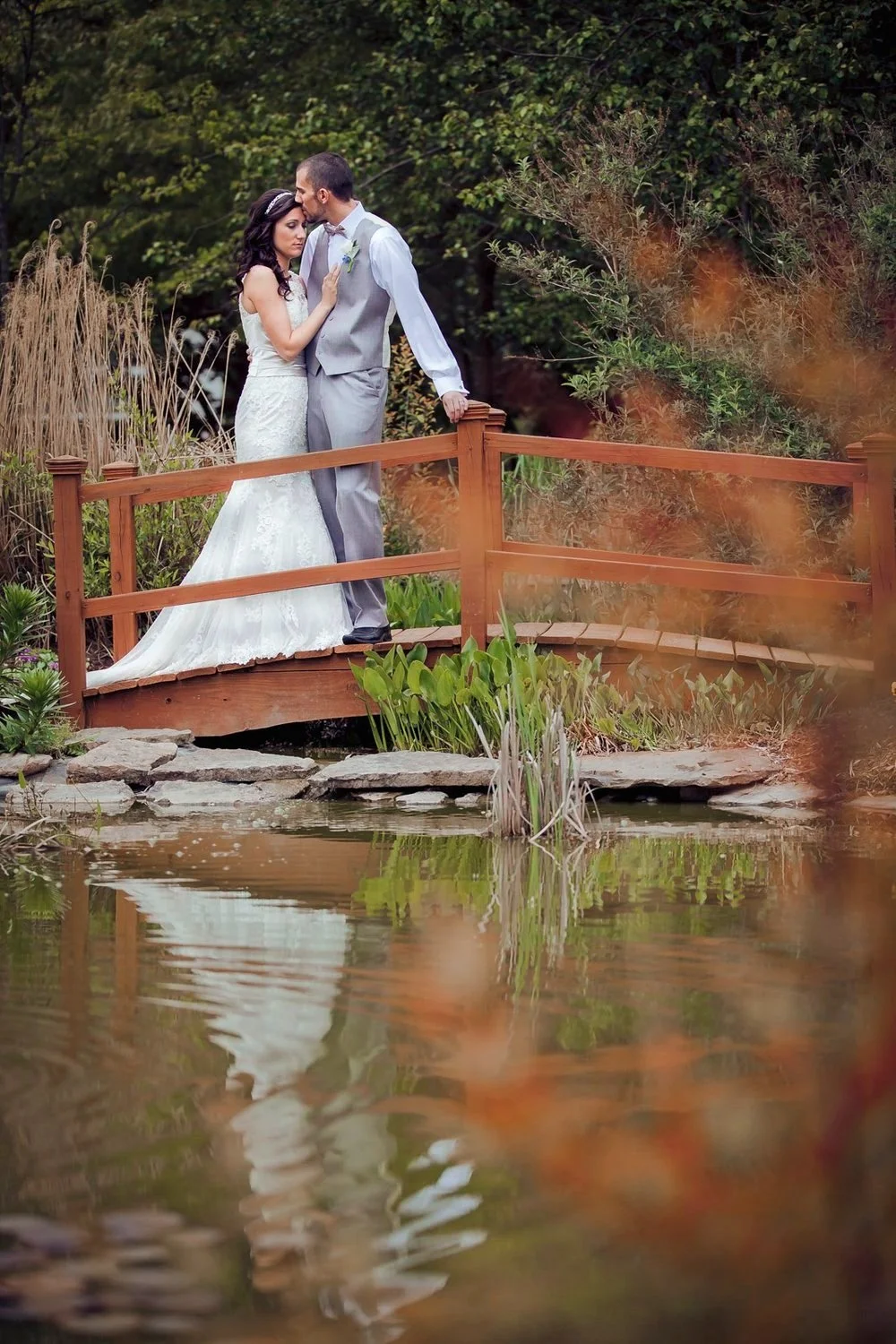 A bride and groom embracing while on a red bridge over a stream.
