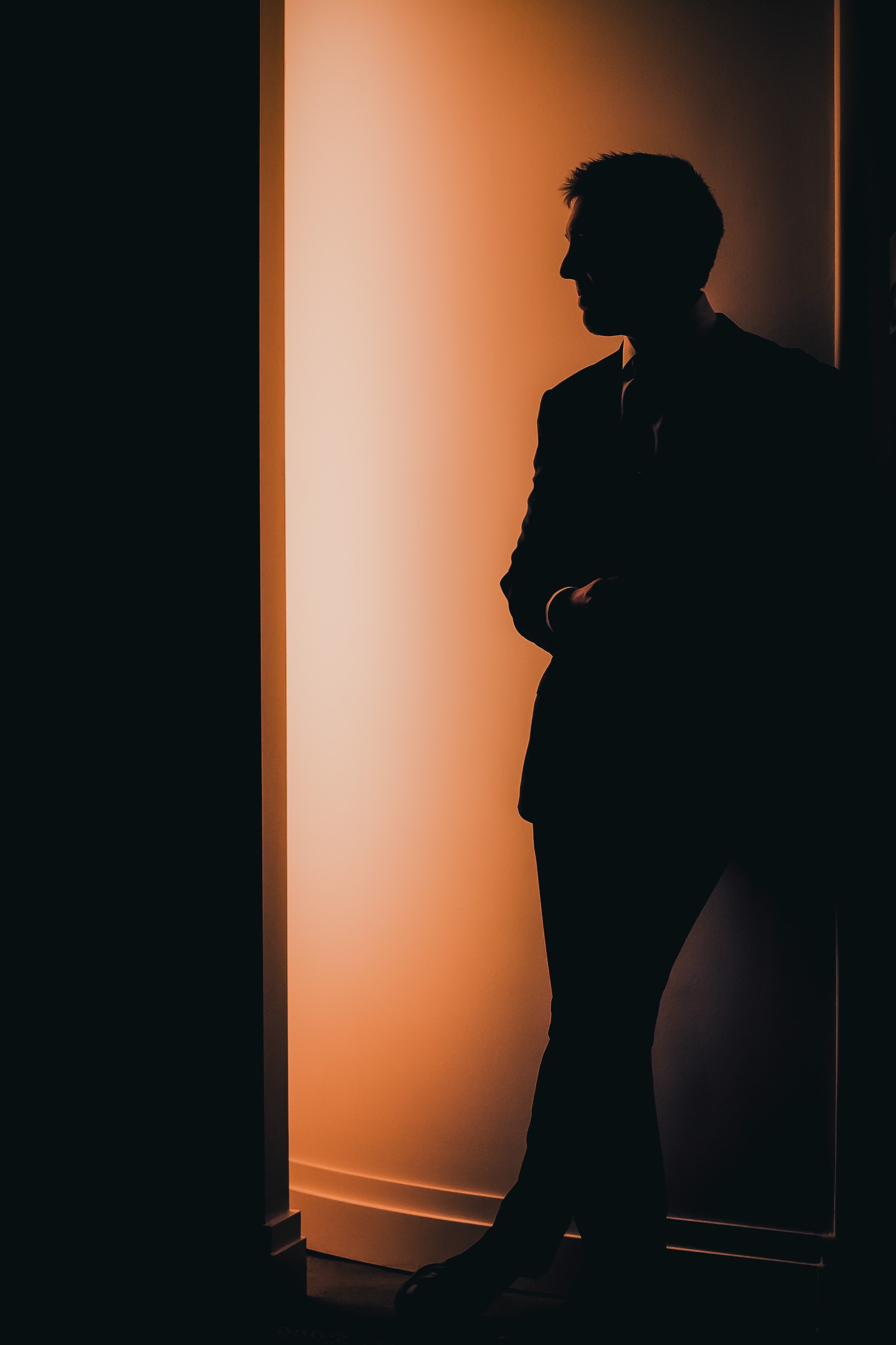A silhouette of a man in a suit standing in front of a backlit wall with warm orange lighting, arms crossed.