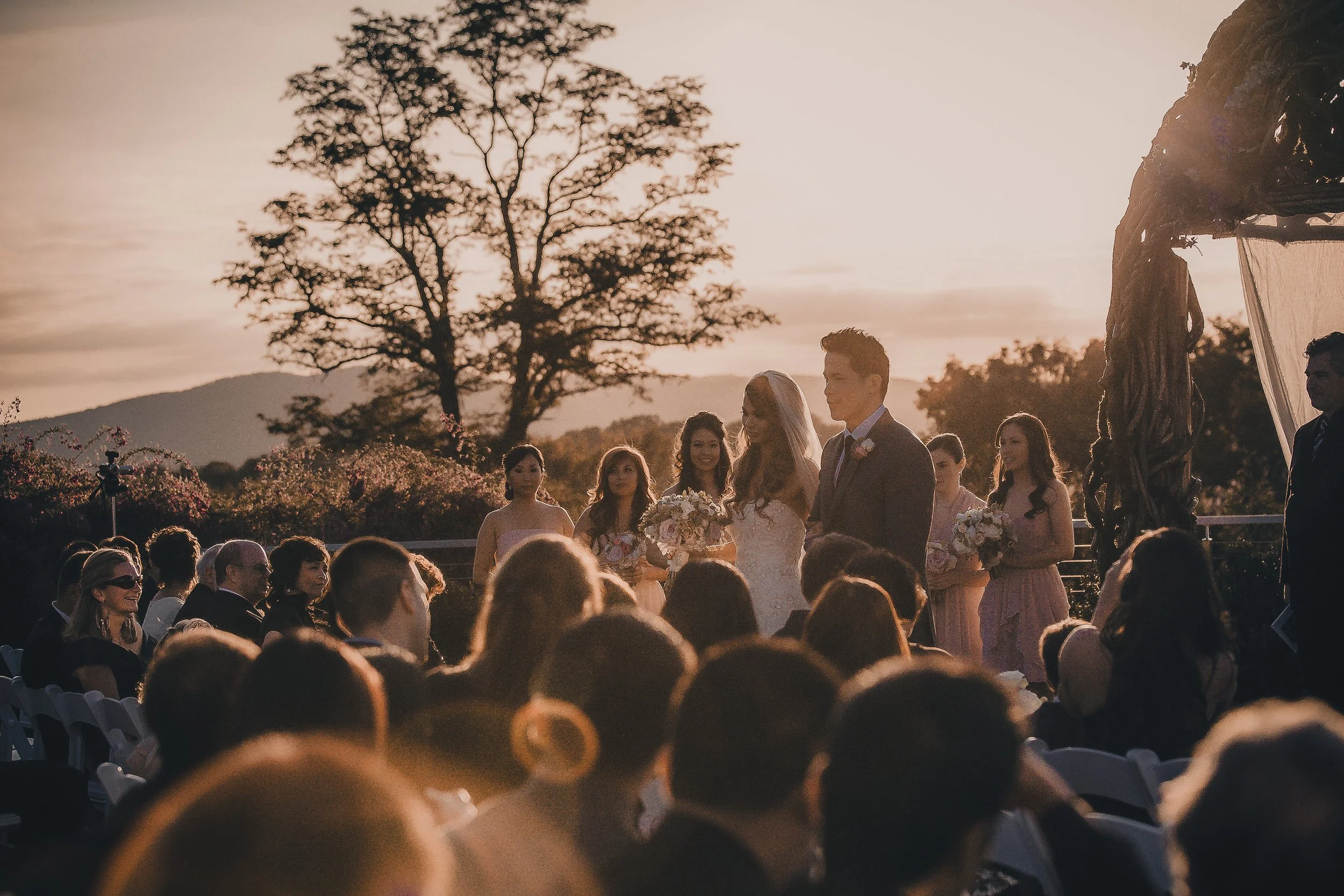 A wedding ceremony outdoors during sunset, with the bride and groom standing facing each other, surrounded by bridesmaids and guests, with large trees and mountains in the background.
