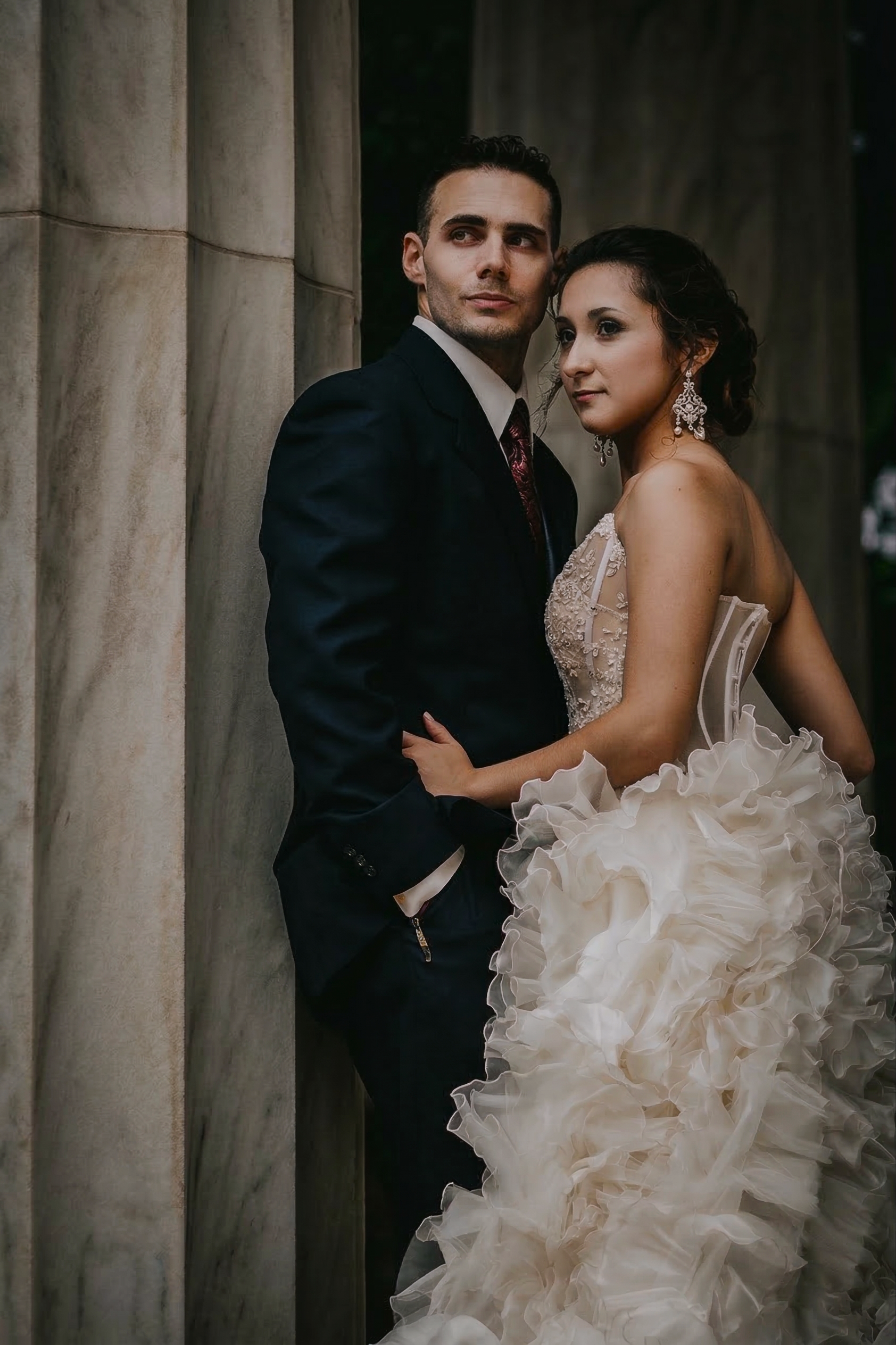 A man and woman dressed in formal attire, standing close together outdoors near stone columns.