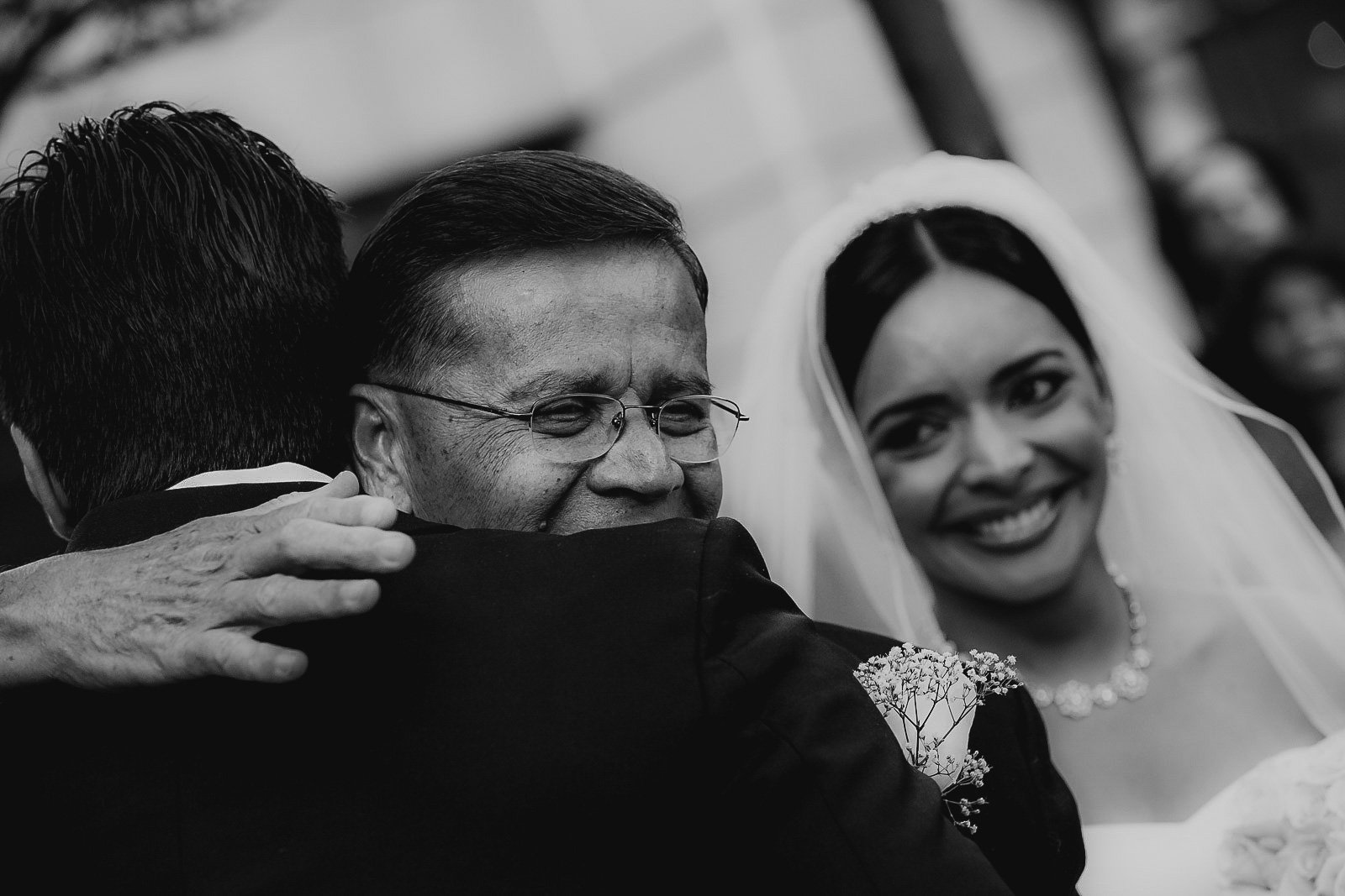 A smiling bride with dark hair and traditional white wedding dress is hugging an older man with glasses and dark hair, who is also hugging another man who is mostly out of the frame.