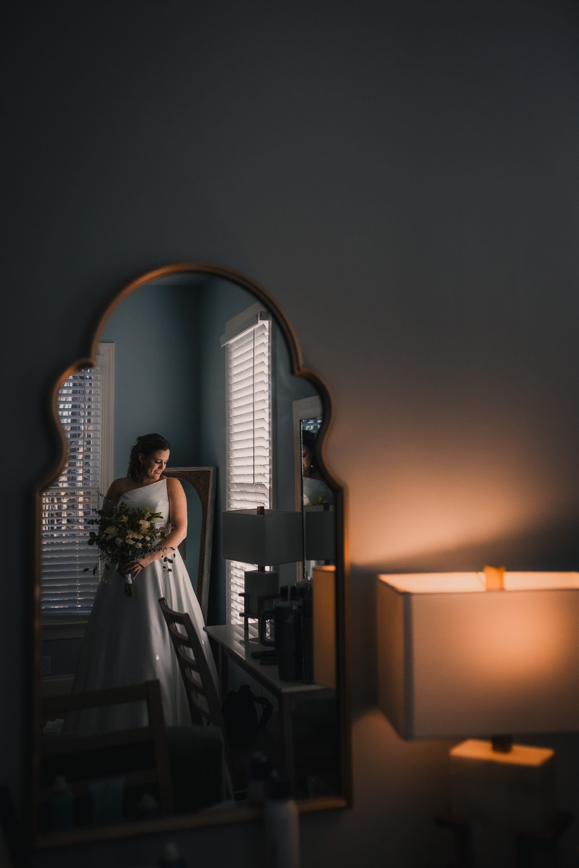 A bride in a white wedding dress holding a bouquet, reflected in a decorative mirror in a softly lit room.