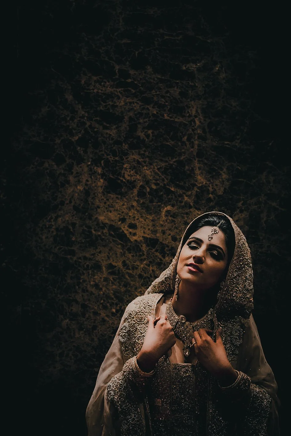 A woman dressed in traditional South Asian bridal attire, wearing ornate jewelry, posing against a dark marble background.
