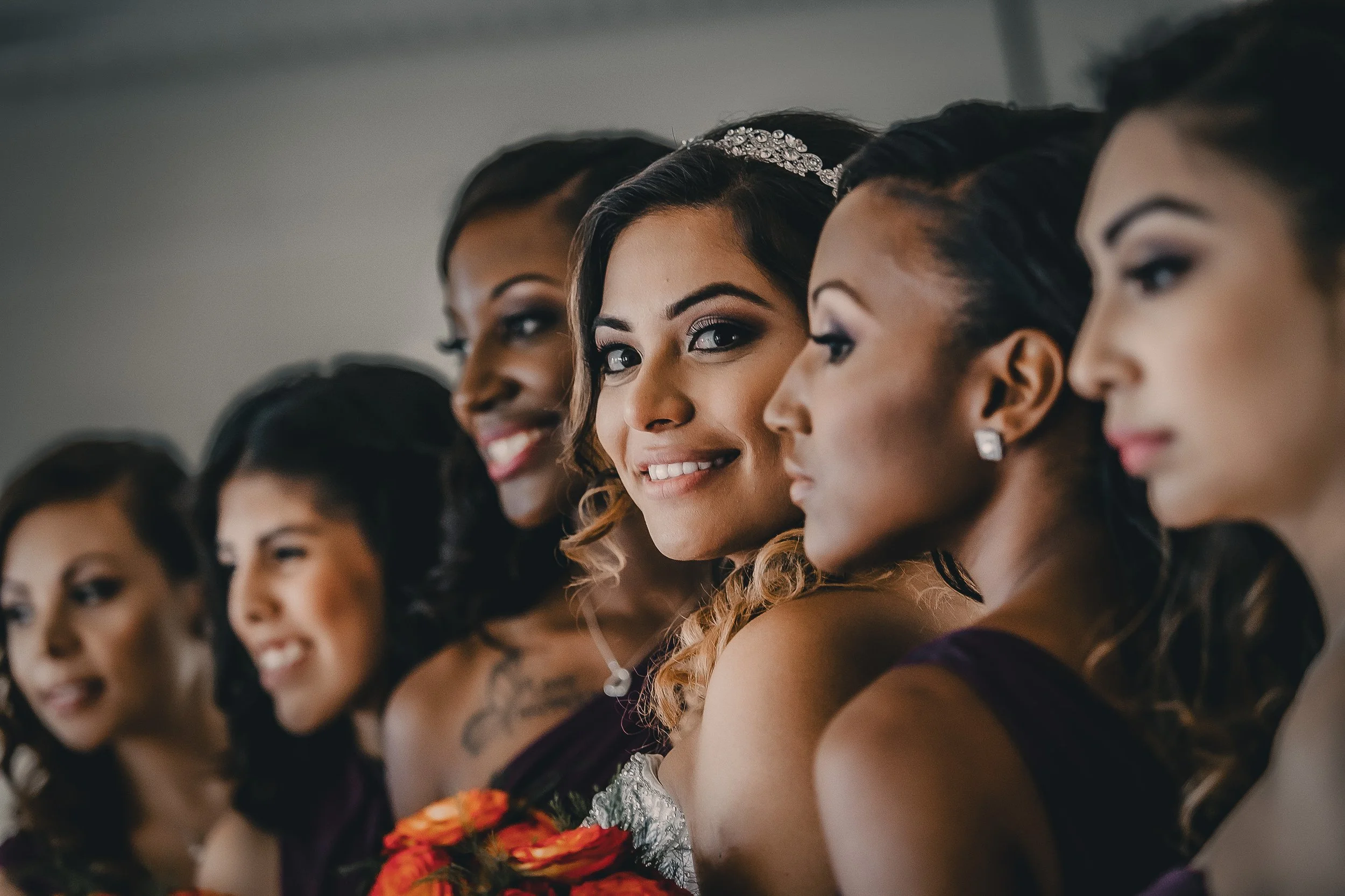 Group of diverse women, including a bride, smiling and looking to the side, dressed in earrings and formal attire at a wedding or celebration.