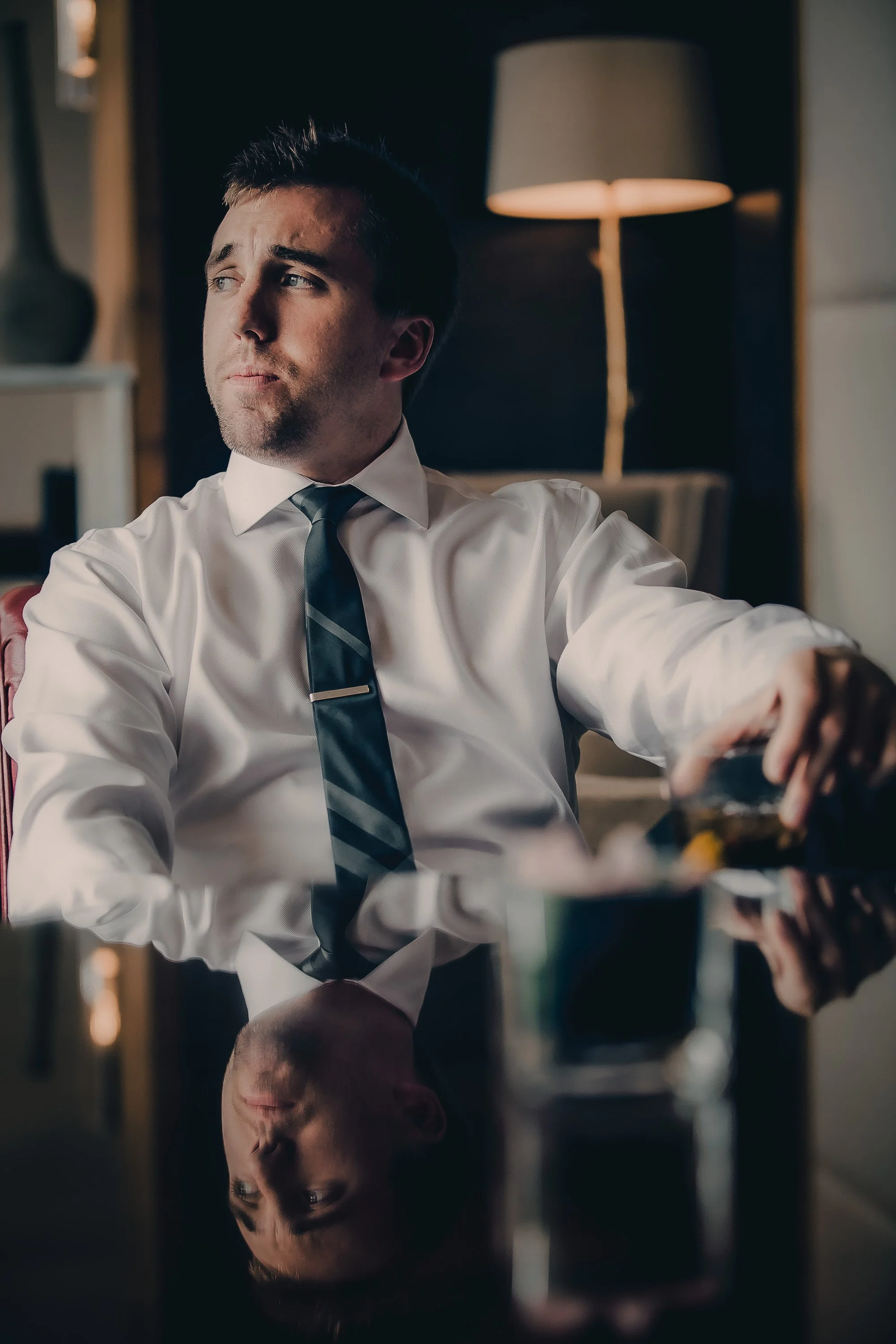 A young man in a white dress shirt and striped tie sitting at a reflective black table, pouring a drink, with a lamp in the background.
