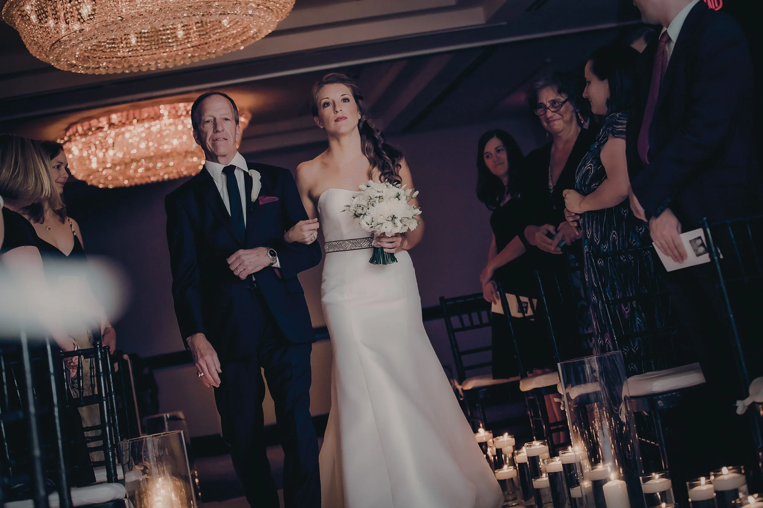 A bride holding a bouquet of white flowers walking down the aisle with her father at her wedding ceremony, surrounded by seated guests in a dimly lit venue with chandeliers and candles.