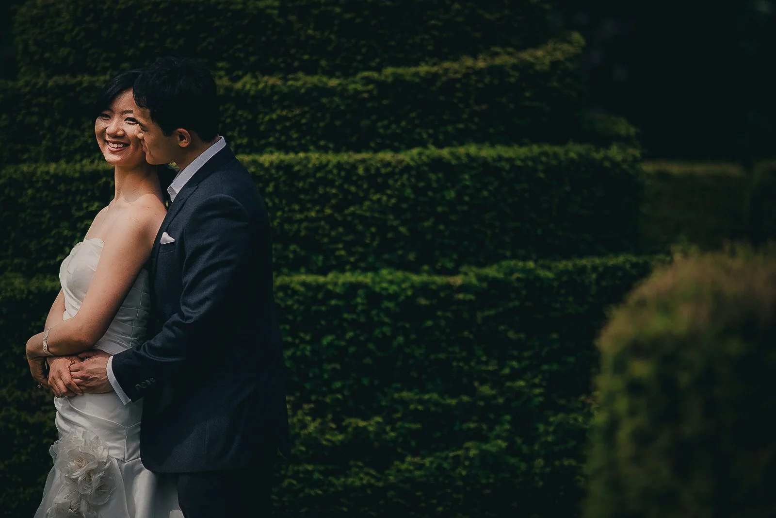 A couple in wedding attire standing close against a background of green bushes, smiling and embracing.
