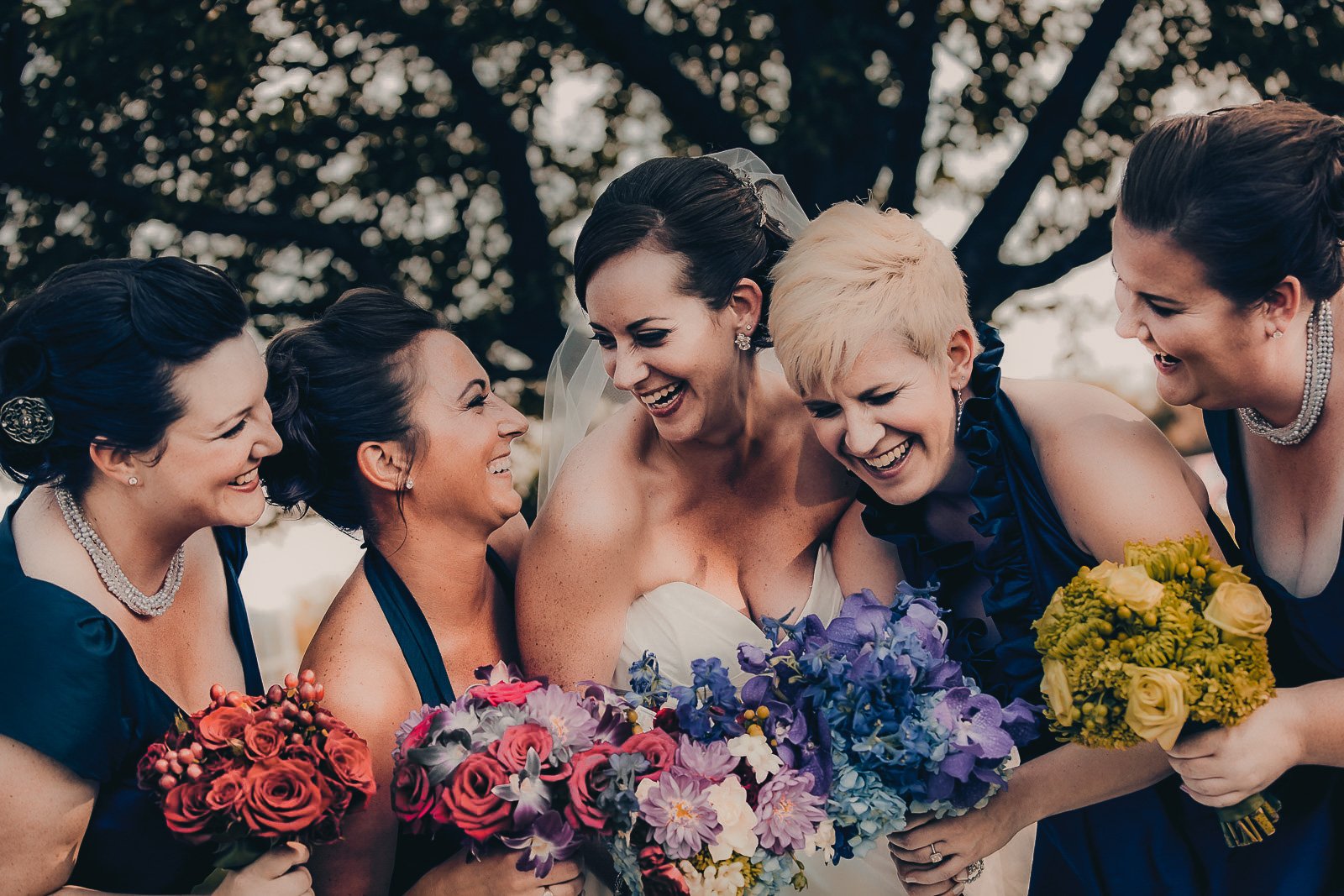 A bride and five bridesmaids smiling and laughing together, holding bouquets of colorful flowers outdoors.