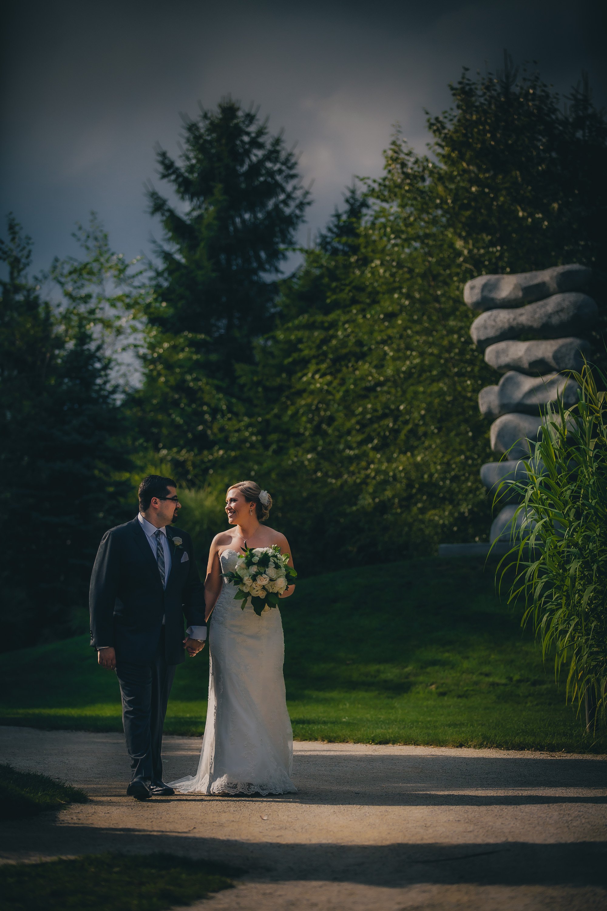 A bride and groom holding hands and walking outdoors, smiling and looking at each other, with wedding attire, a lush green background, and rock formations in the distance.
