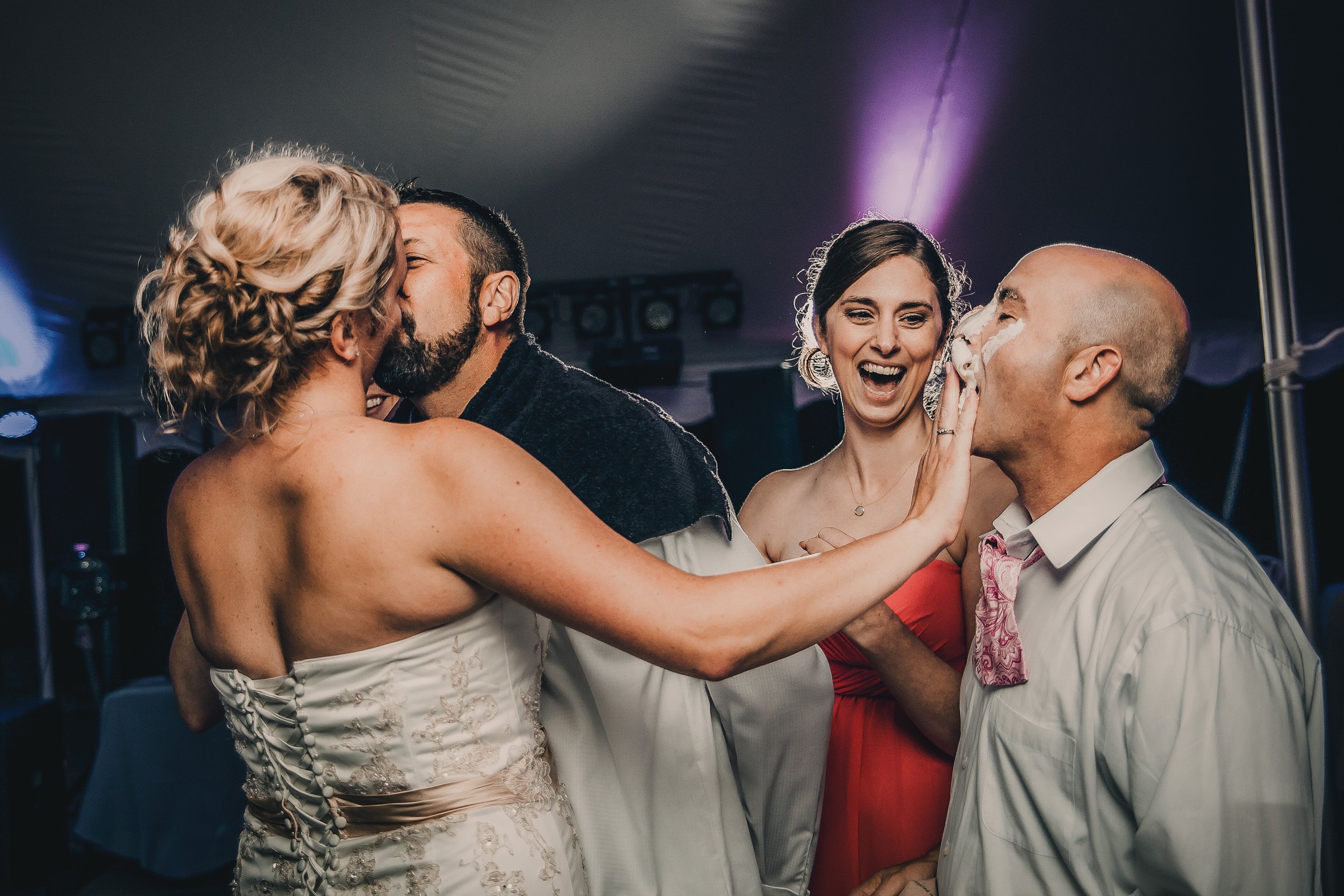 A wedding celebration with a bride and groom kissing, surrounded by two women, one laughing and touching the groom's face, and the other smiling, in a dimly lit venue.
