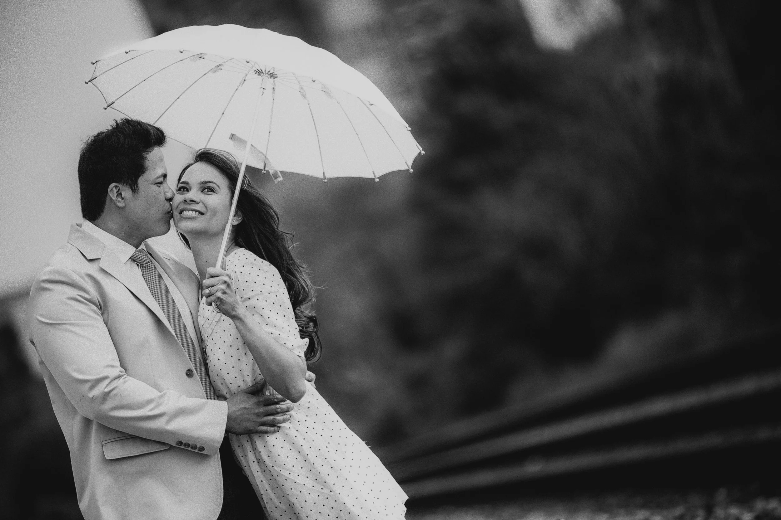 A couple in formal attire under a white umbrella, with the man kissing the woman on the cheek. The woman smiles, holding the umbrella handle, and they are outdoors in a natural setting.