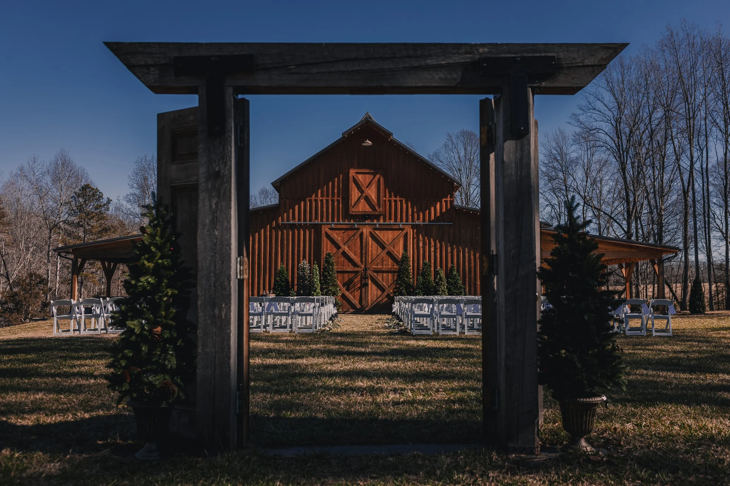 A rustic barn decorated for a wedding with Christmassy pine trees and white chairs arranged outside, viewed through a wooden archway with trees in the background.