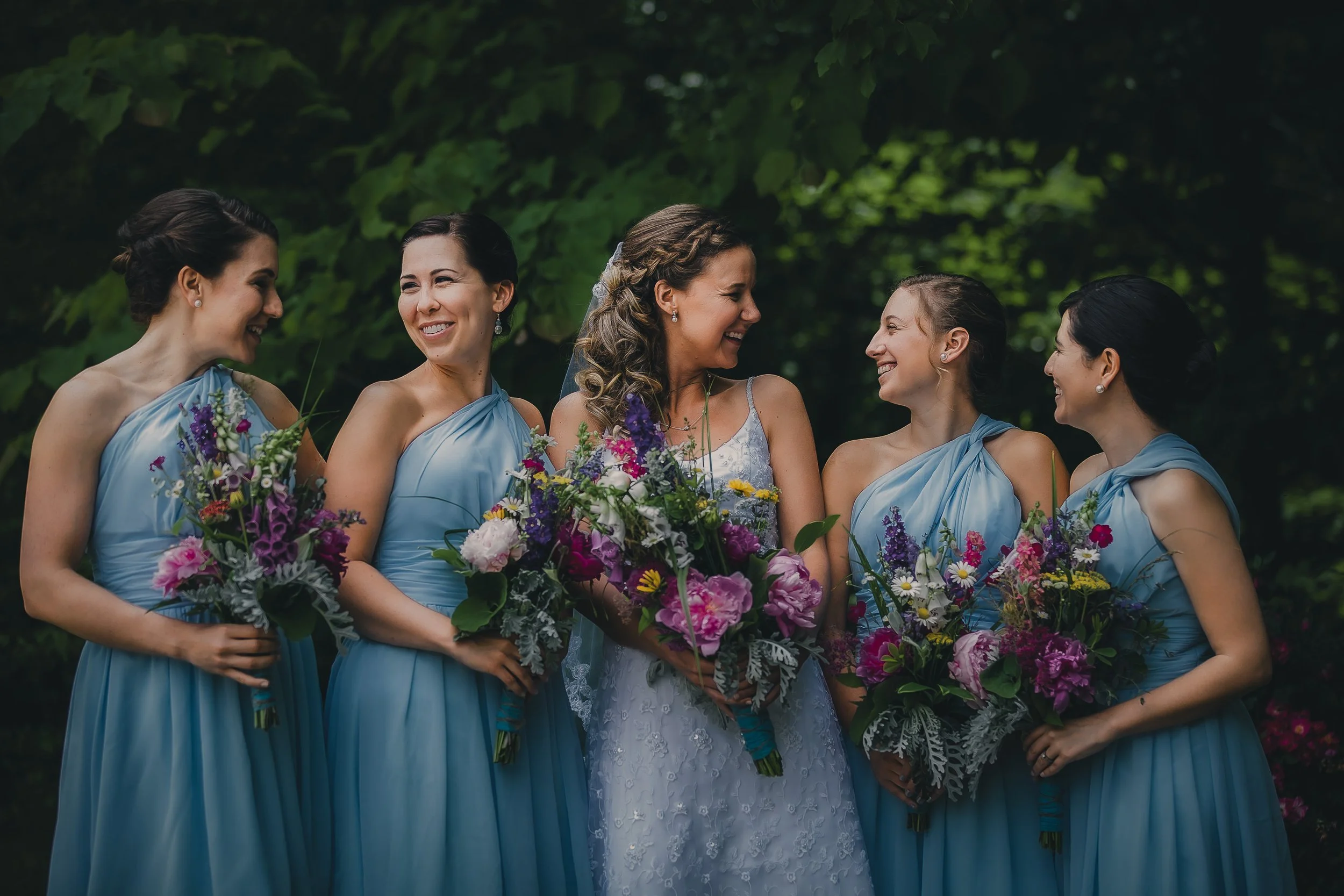 Bride and five bridesmaids in blue dresses, holding bouquets of flowers, smiling and enjoying each other's company outdoors in a green, leafy setting.