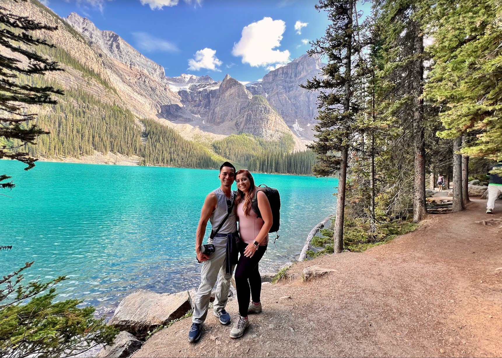 A couple standing on a dirt path beside a turquoise lake with mountains and pine trees in the background.