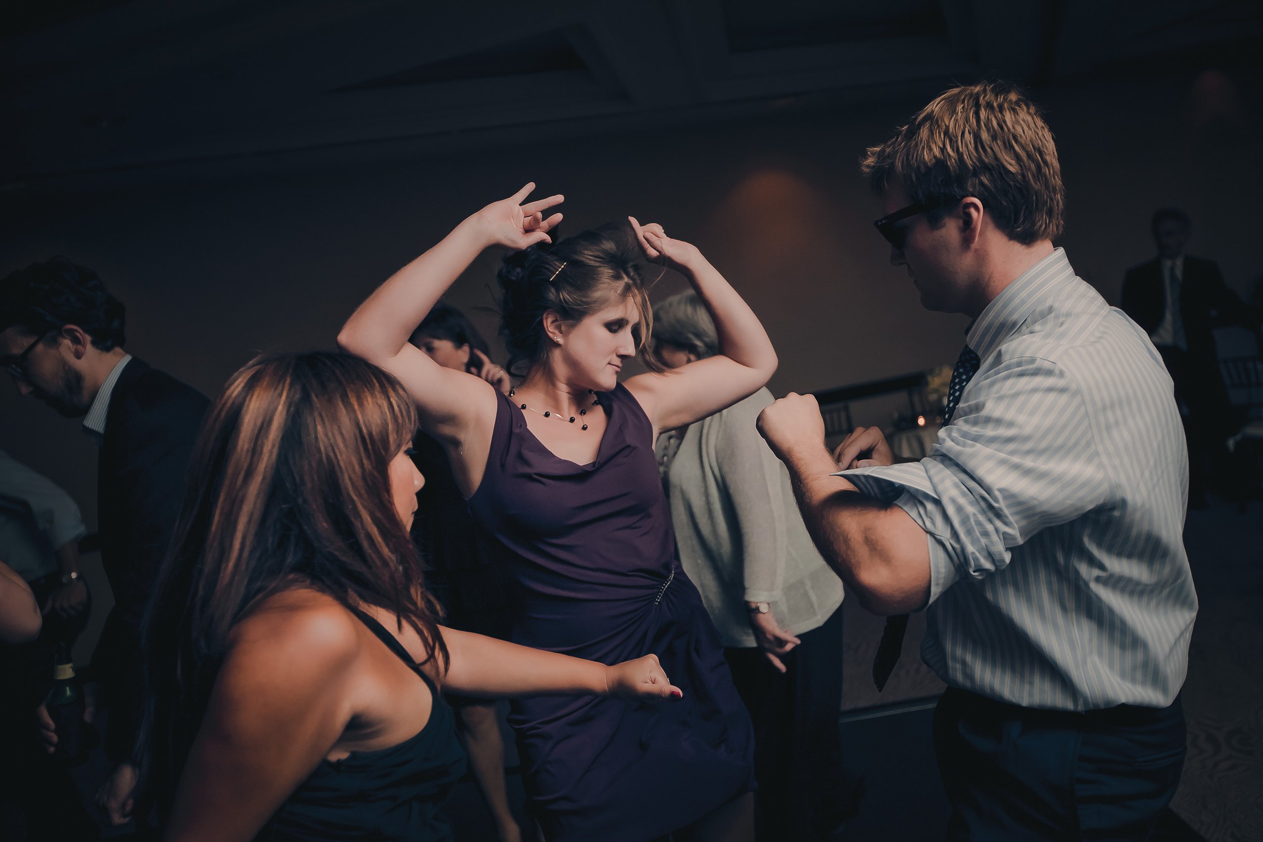 People dancing at an indoor party, with a woman in a purple dress and a man in a striped shirt in the foreground.