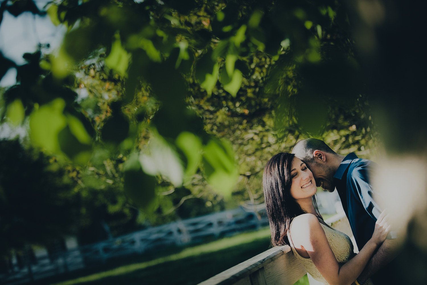A couple sharing a joyful moment under green leafy trees on a sunny day, with a woman smiling and a man leaning in close.
